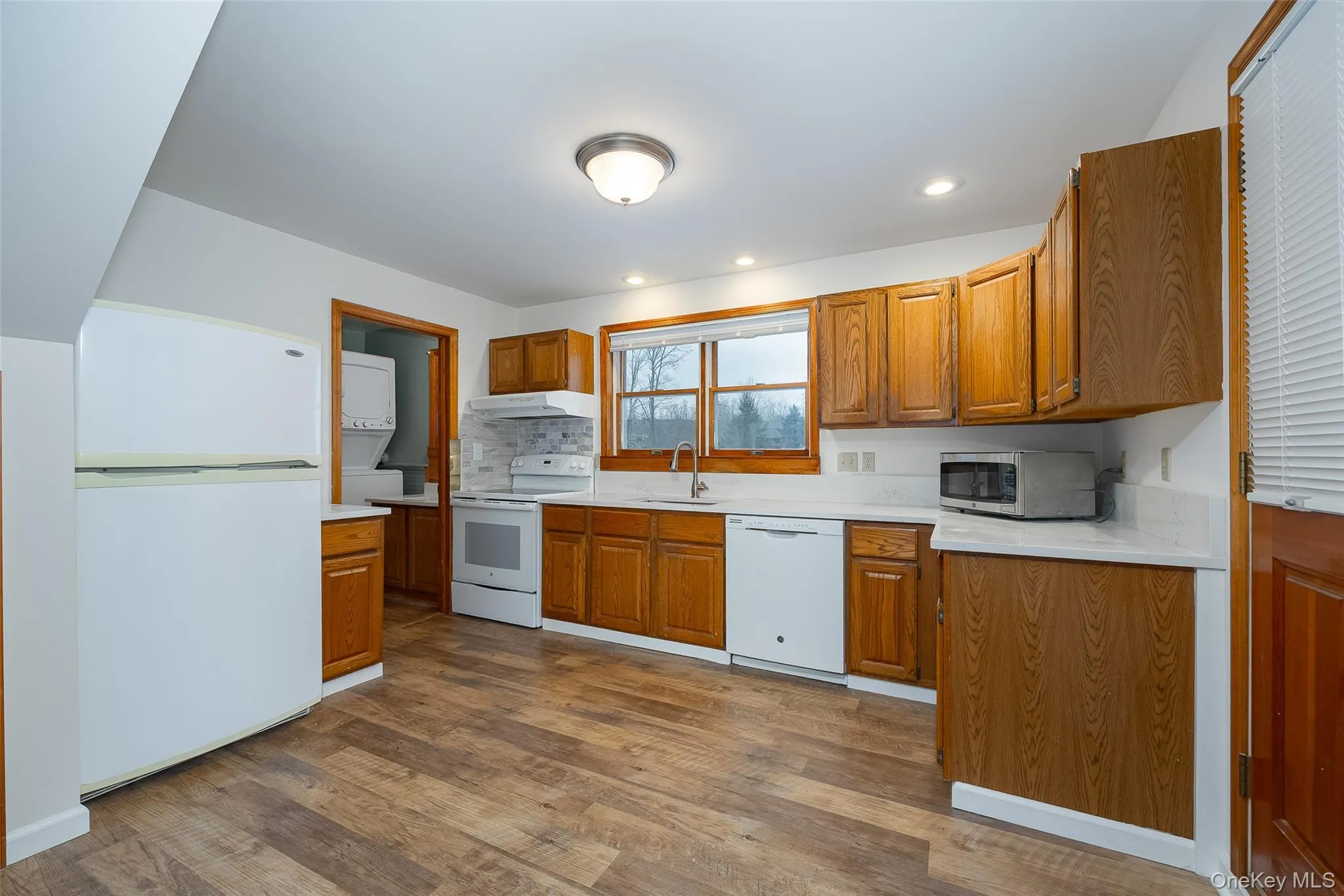 Kitchen with white appliances, stacked washer and clothes dryer, brown cabinetry, dark wood finished floors, and recessed lighting #2 Kitchen with white appliances, stacked washer and clothes dryer, brown cabinetry, dark wood finished floors, and recessed lighting #2