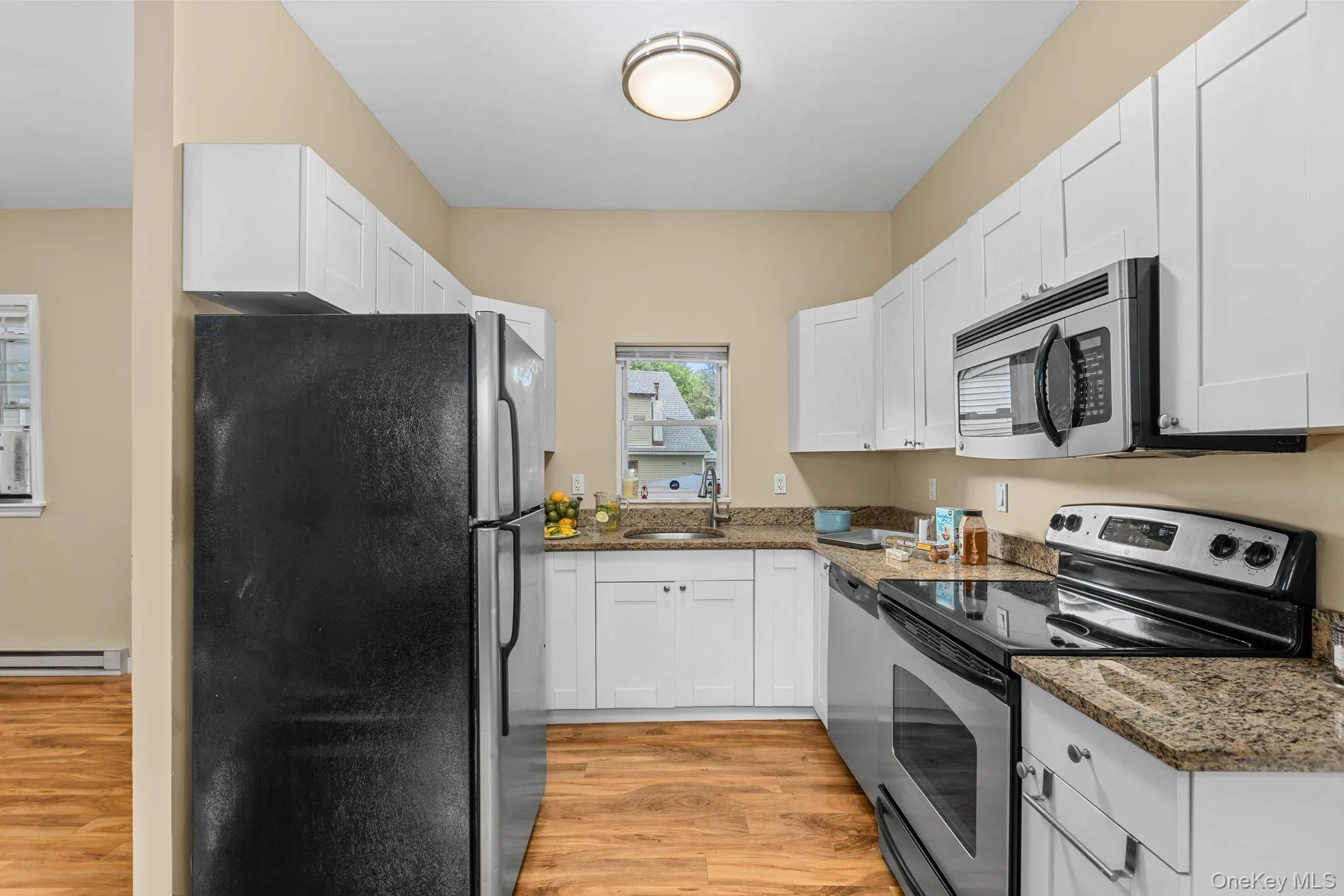Galley kitchen featuring white soft-close cabinetry, wood-finish flooring, stainless steel appliances, granite-style countertops, and a ceiling-mounted light fixture. Galley kitchen featuring white soft-close cabinetry, wood-finish flooring, stainless steel appliances, granite-style countertops, and a ceiling-mounted light fixture.