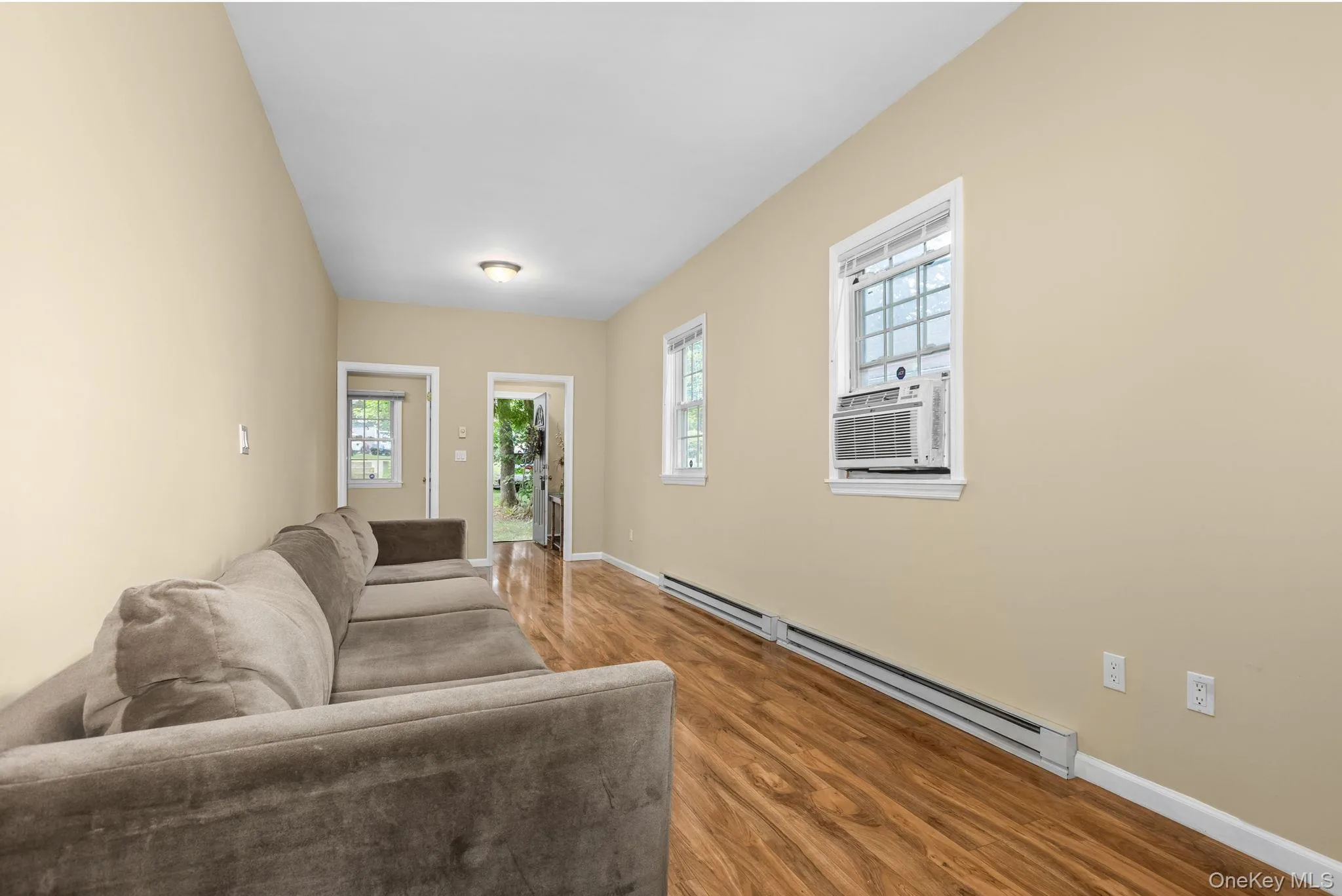 Spacious room featuring wood-finish flooring, two windows with white trim, a window-mounted air conditioning unit, and baseboard heating Spacious room featuring wood-finish flooring, two windows with white trim, a window-mounted air conditioning unit, and baseboard heating