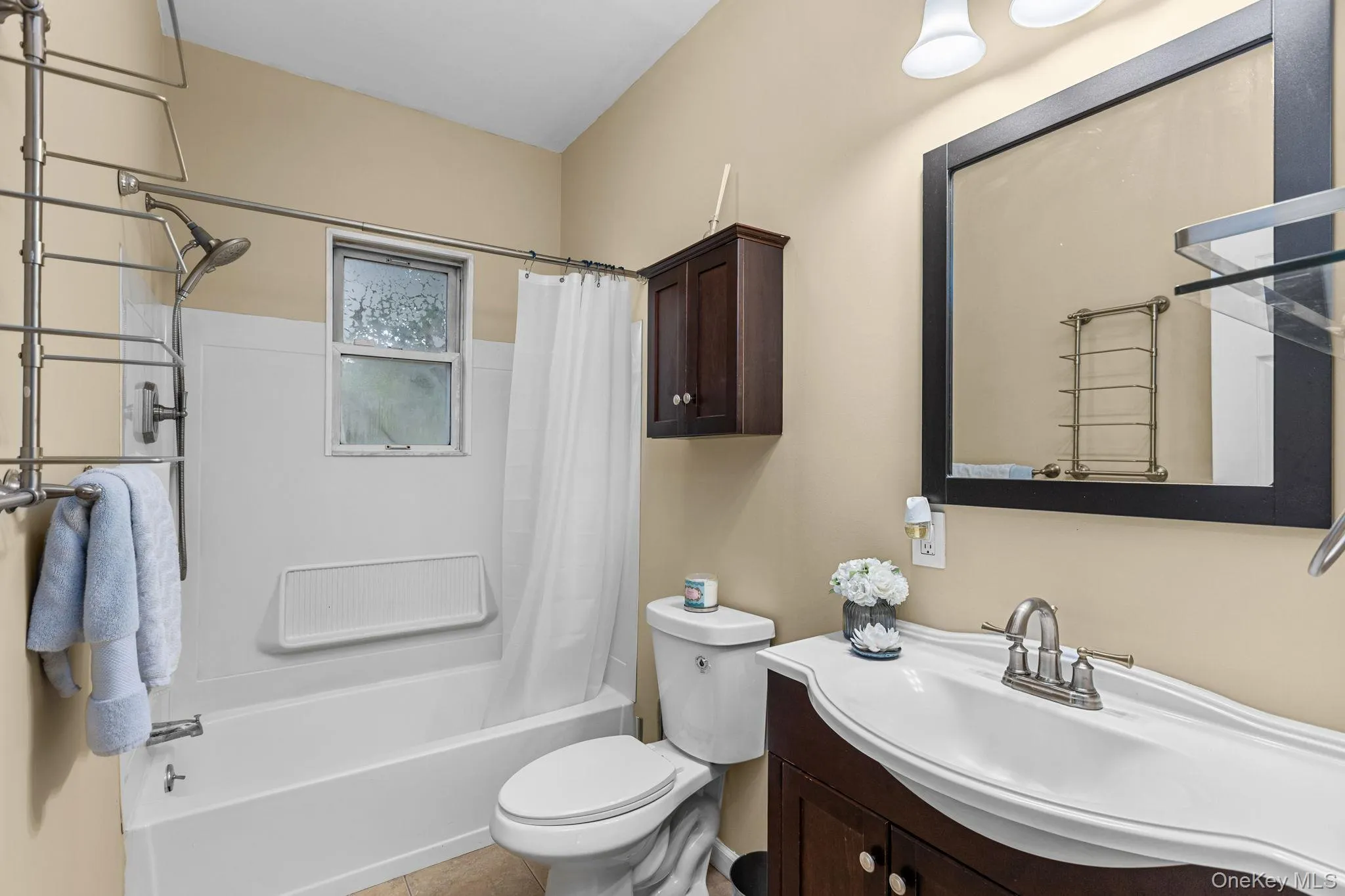 Bathroom featuring a tub/shower combination with a window, a dark wood-finish vanity with an integrated white sink, a matching framed mirror, and a wall-mounted wood-finish cabinet Bathroom featuring a tub/shower combination with a window, a dark wood-finish vanity with an integrated white sink, a matching framed mirror, and a wall-mounted wood-finish cabinet