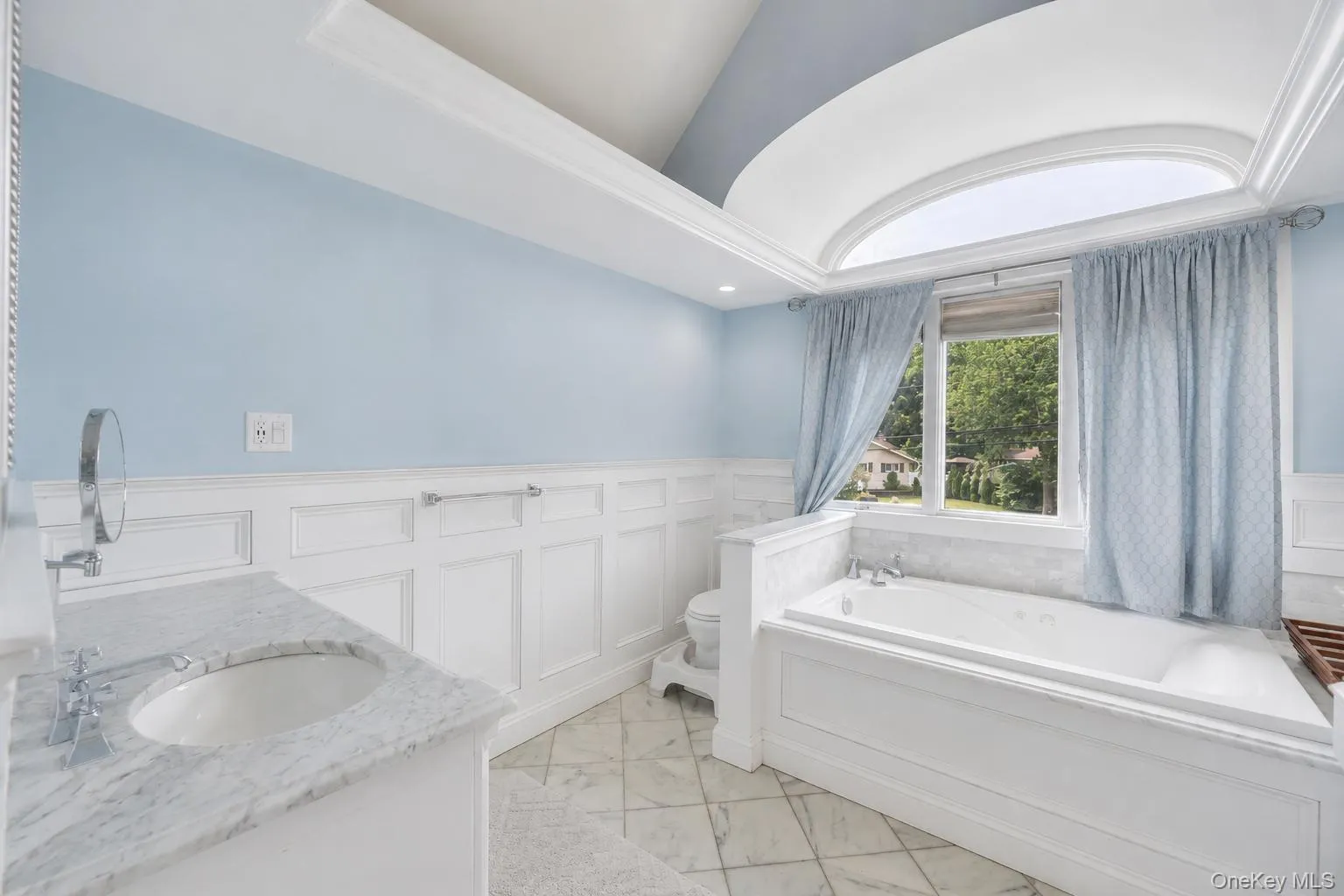 Bathroom featuring a spacious built-in soaking tub, an arched transom window, a single vanity with a stone countertop and undermount sink, wainscoting, and large format floor tiles Bathroom featuring a spacious built-in soaking tub, an arched transom window, a single vanity with a stone countertop and undermount sink, wainscoting, and large format floor tiles