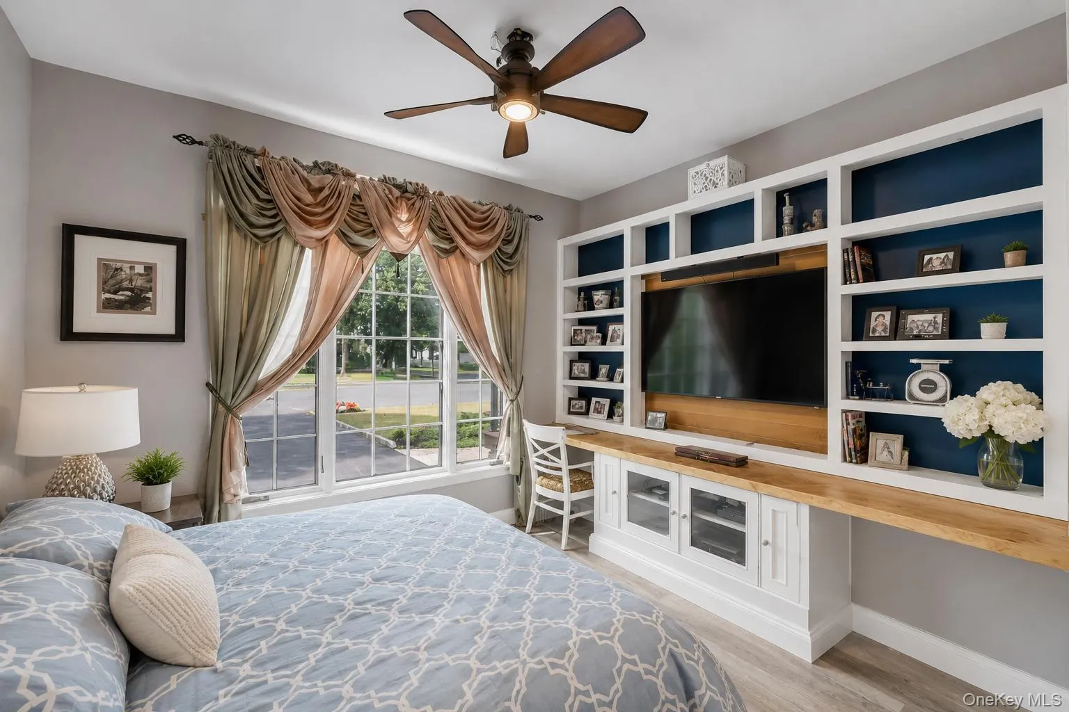 Room featuring a large window with draped valances, a ceiling fan with integrated lighting, and wood-finish flooring Room featuring a large window with draped valances, a ceiling fan with integrated lighting, and wood-finish flooring