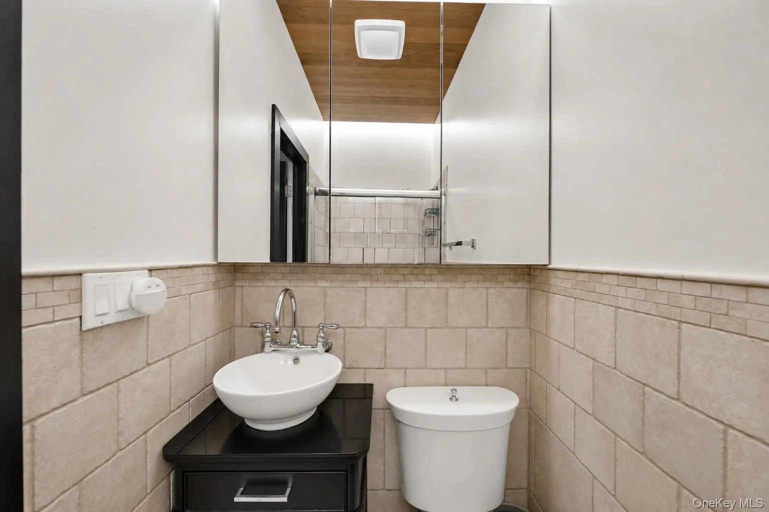 Bathroom featuring a vessel sink with chrome fixtures, a dark wood-finish vanity with drawer, and light-toned tiled walls with decorative banding Bathroom featuring a vessel sink with chrome fixtures, a dark wood-finish vanity with drawer, and light-toned tiled walls with decorative banding