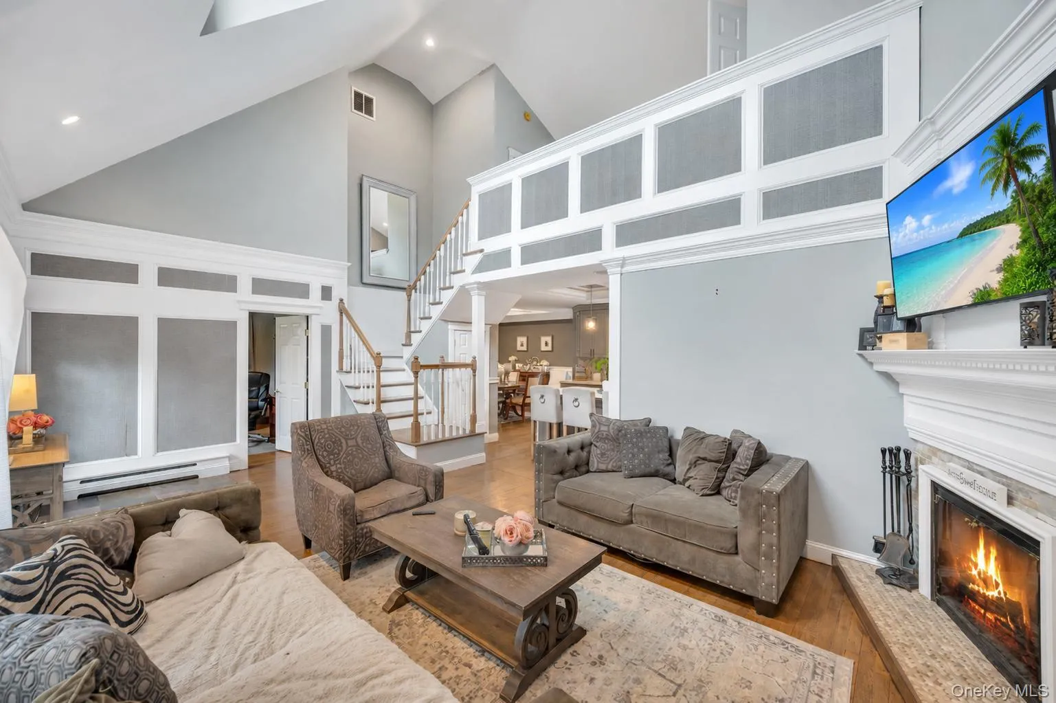 Two-story living area featuring a vaulted ceiling with skylights, hardwood-finish flooring, a fireplace with a white mantel, and an open staircase with wood railings Two-story living area featuring a vaulted ceiling with skylights, hardwood-finish flooring, a fireplace with a white mantel, and an open staircase with wood railings