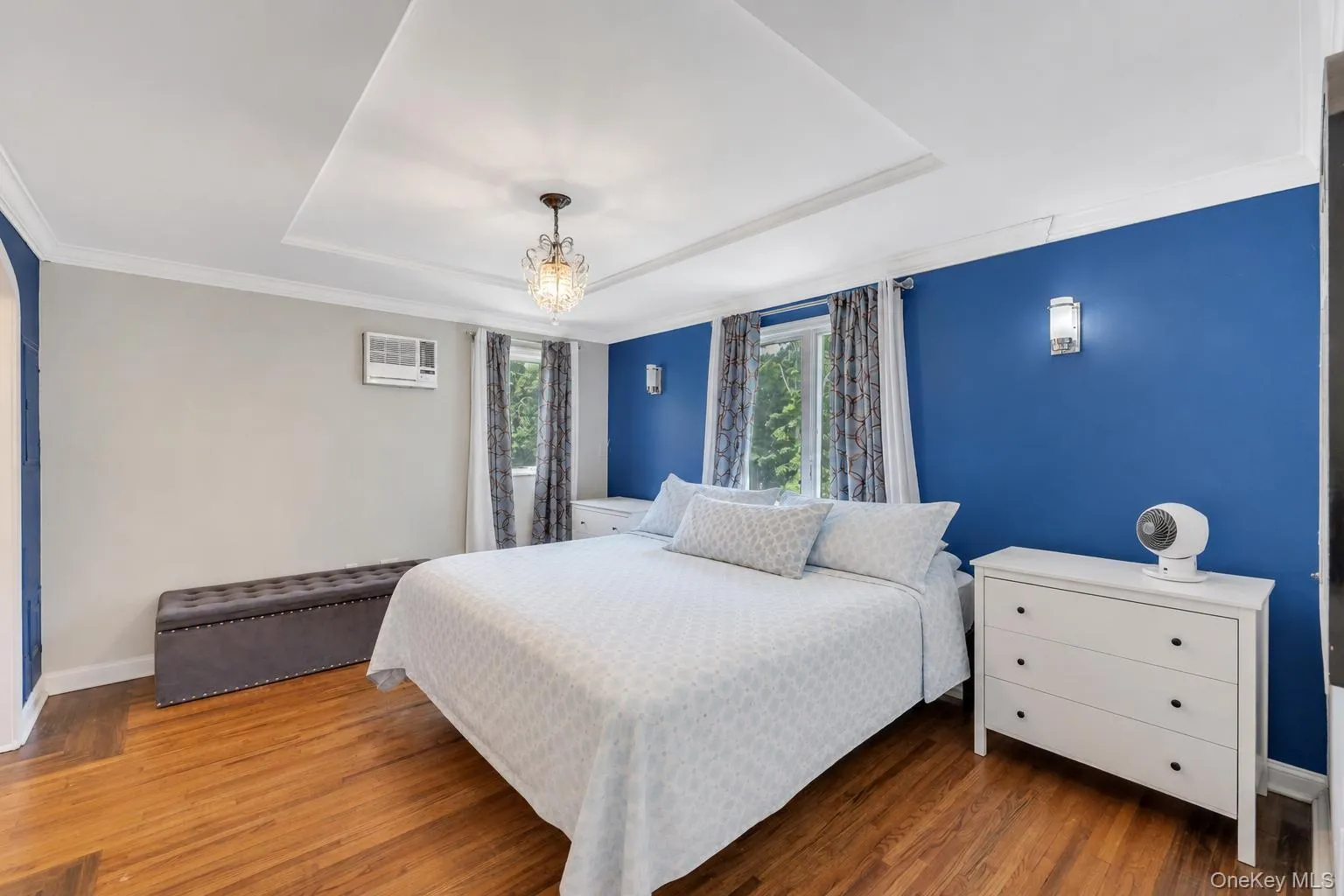 Bedroom featuring wood-finish flooring, a tray ceiling with a chandelier, crown molding, two windows with curtain rods, and two wall sconces Bedroom featuring wood-finish flooring, a tray ceiling with a chandelier, crown molding, two windows with curtain rods, and two wall sconces