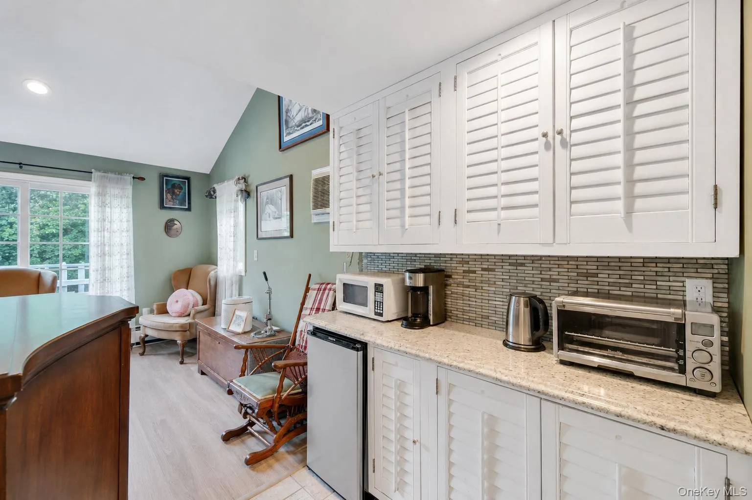 Galley area featuring white shaker-style cabinetry with louvered doors, a mosaic tile backsplash, and a granite-finish countertop Galley area featuring white shaker-style cabinetry with louvered doors, a mosaic tile backsplash, and a granite-finish countertop