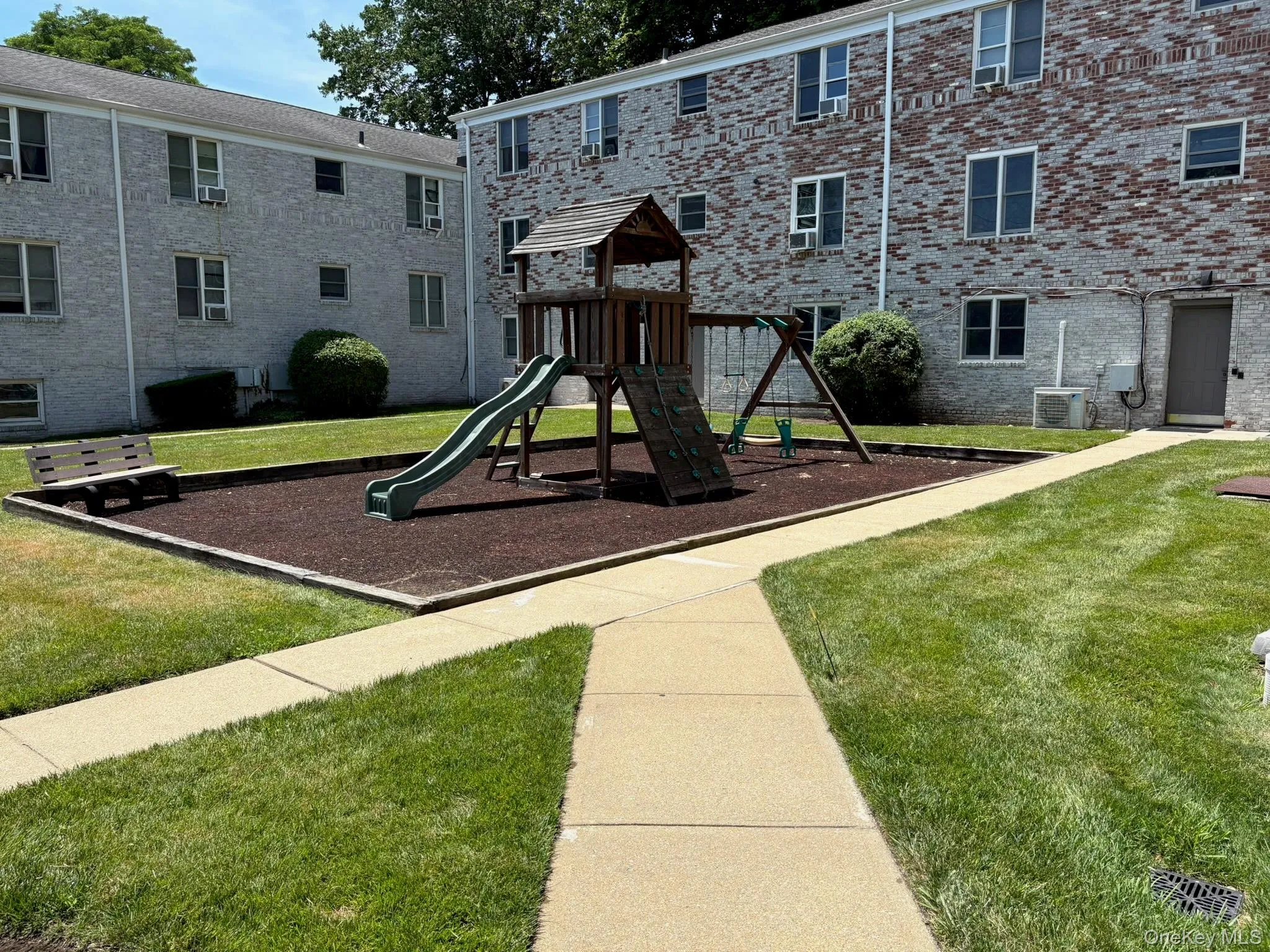 Outdoor recreation area featuring a multi-level playset with a slide and swings, bordered by a concrete walkway Outdoor recreation area featuring a multi-level playset with a slide and swings, bordered by a concrete walkway