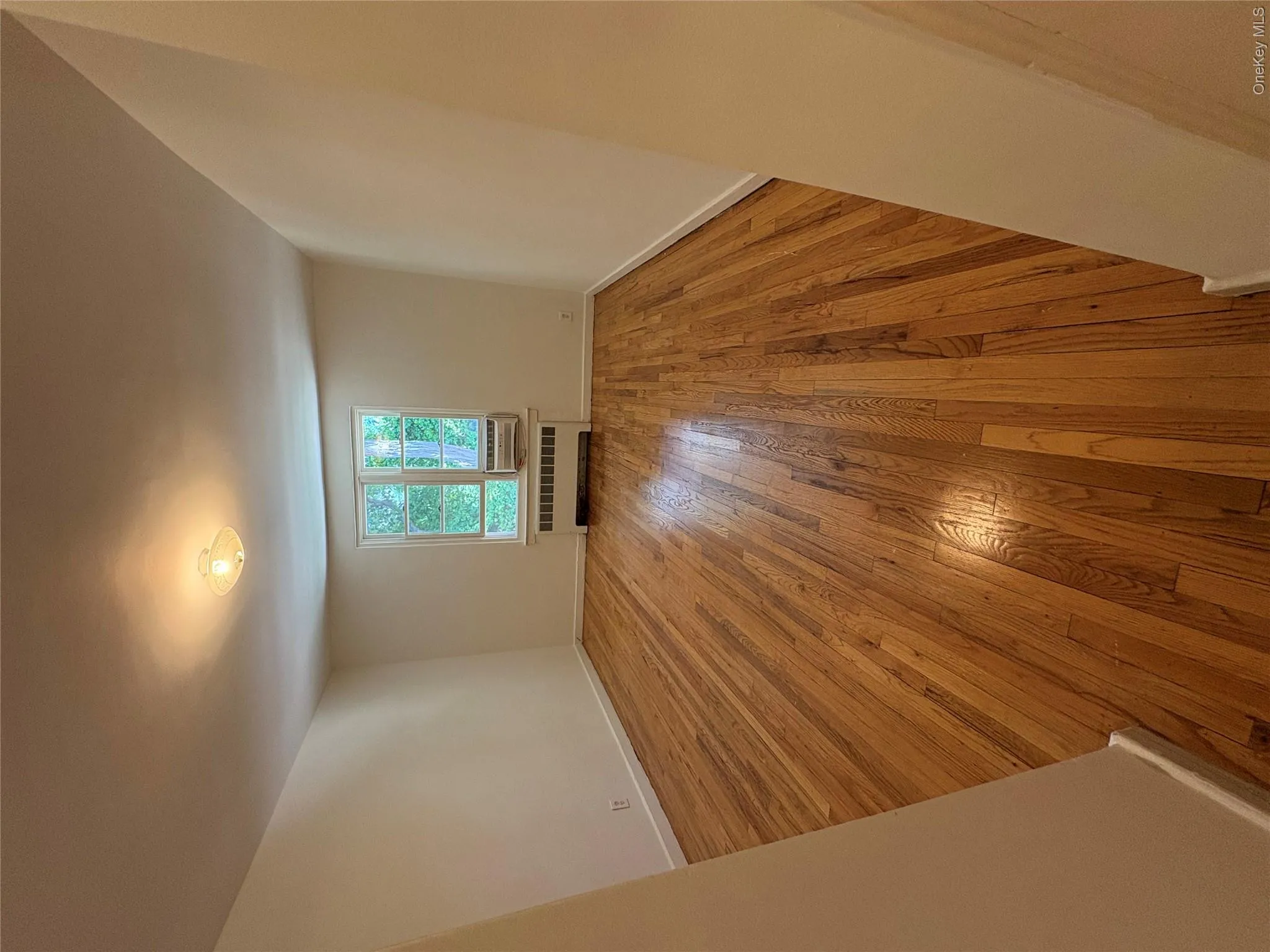 Spacious room featuring wood-finish flooring, a window with exterior views, an air conditioning unit, and a ceiling-mounted light fixture. View from hallway. Spacious room featuring wood-finish flooring, a window with exterior views, an air conditioning unit, and a ceiling-mounted light fixture. View from hallway.