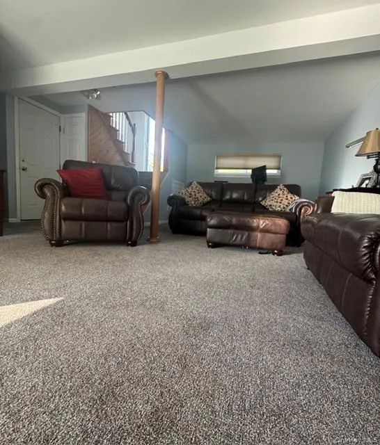 Spacious room featuring neutral carpeting, a staircase with wood-finish railing, and a light-toned ceiling with exposed beam Spacious room featuring neutral carpeting, a staircase with wood-finish railing, and a light-toned ceiling with exposed beam