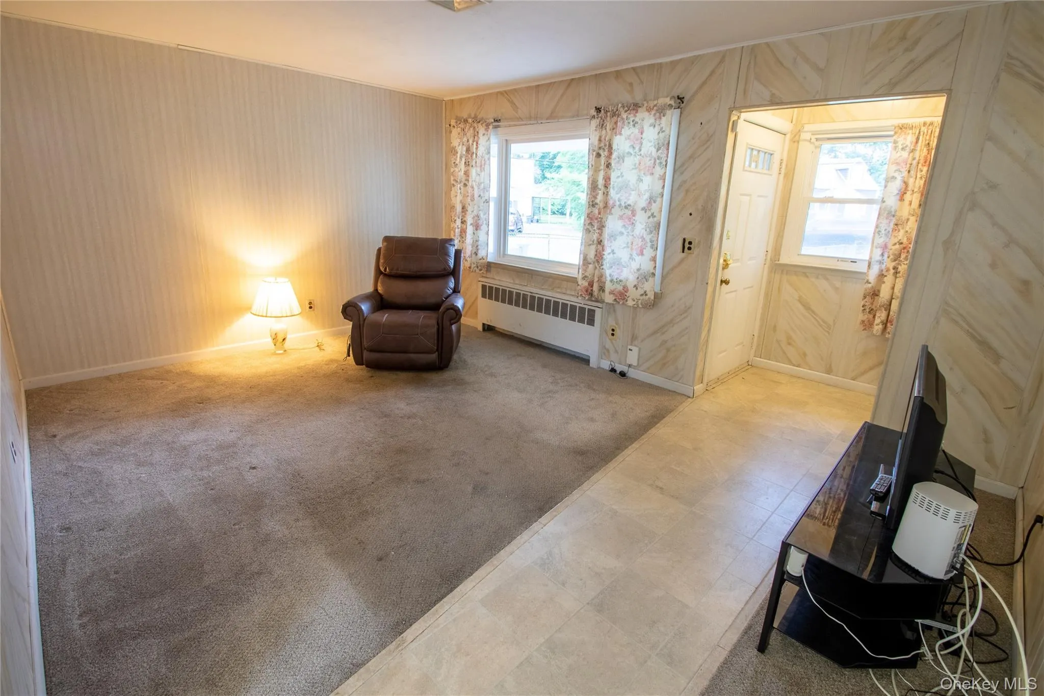 Carpeted living area with textured wallpaper and a doorway featuring paneling, windows with curtains, and light-toned flooring Carpeted living area with textured wallpaper and a doorway featuring paneling, windows with curtains, and light-toned flooring
