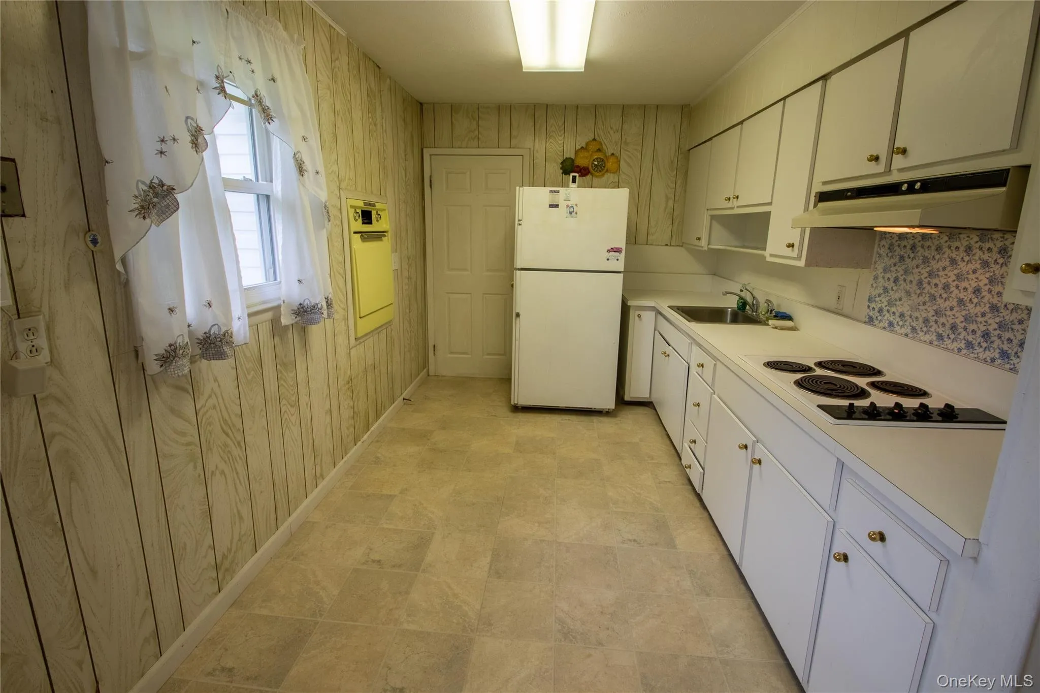 Kitchen featuring light-toned wood-finish paneling, an integrated electric cooktop, white cabinetry with brass-toned hardware, a stainless steel sink, and light-toned tile flooring Kitchen featuring light-toned wood-finish paneling, an integrated electric cooktop, white cabinetry with brass-toned hardware, a stainless steel sink, and light-toned tile flooring