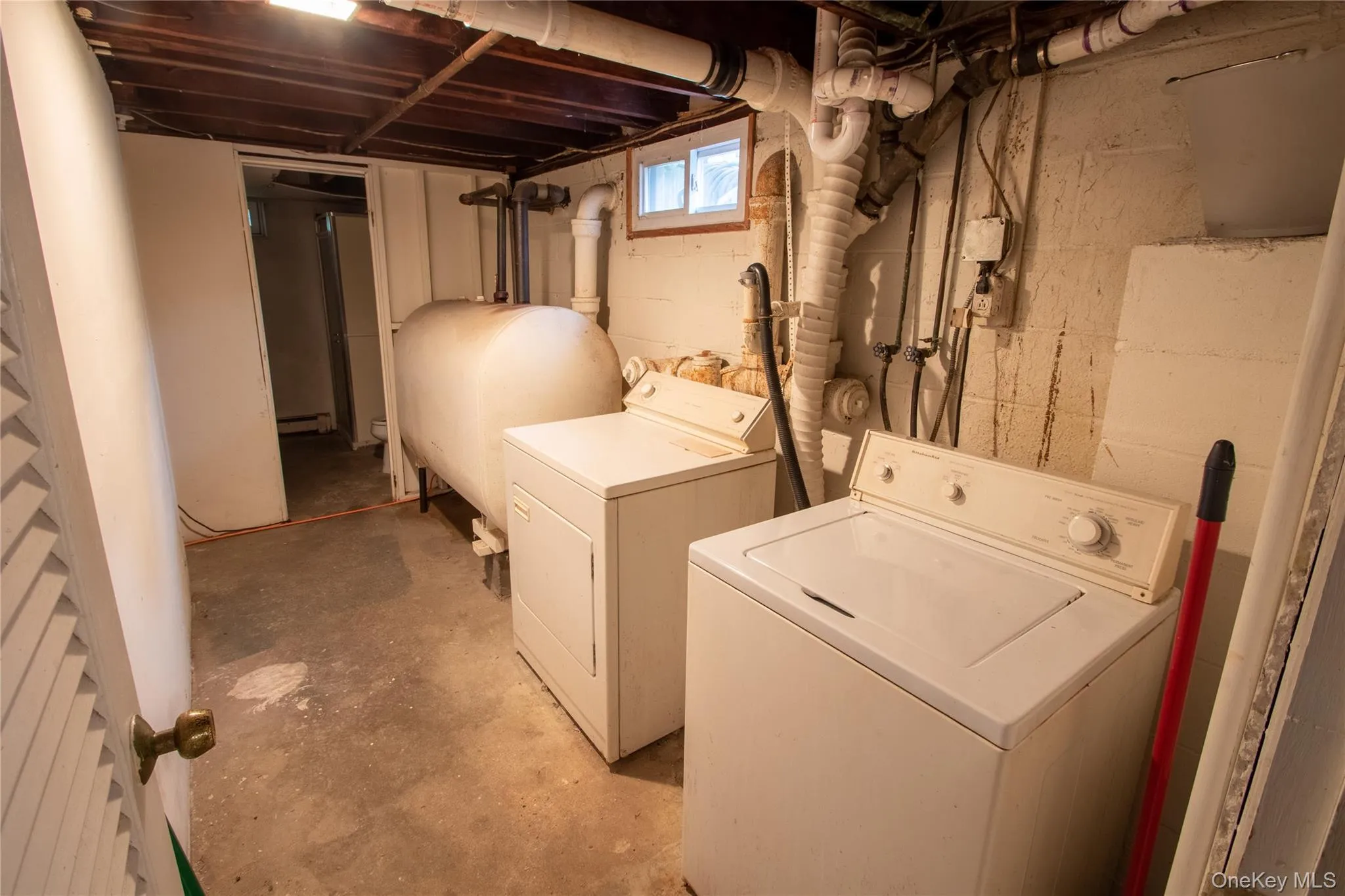 Basement utility area featuring a washer and dryer pair, exposed overhead joists, concrete flooring, and a large fuel tank Basement utility area featuring a washer and dryer pair, exposed overhead joists, concrete flooring, and a large fuel tank