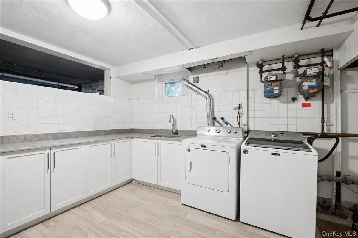 Laundry area featuring wood-finish flooring, white cabinetry with silver hardware, light-toned countertops, a stainless steel sink with a gooseneck faucet, and white subway tile backsplash Laundry area featuring wood-finish flooring, white cabinetry with silver hardware, light-toned countertops, a stainless steel sink with a gooseneck faucet, and white subway tile backsplash