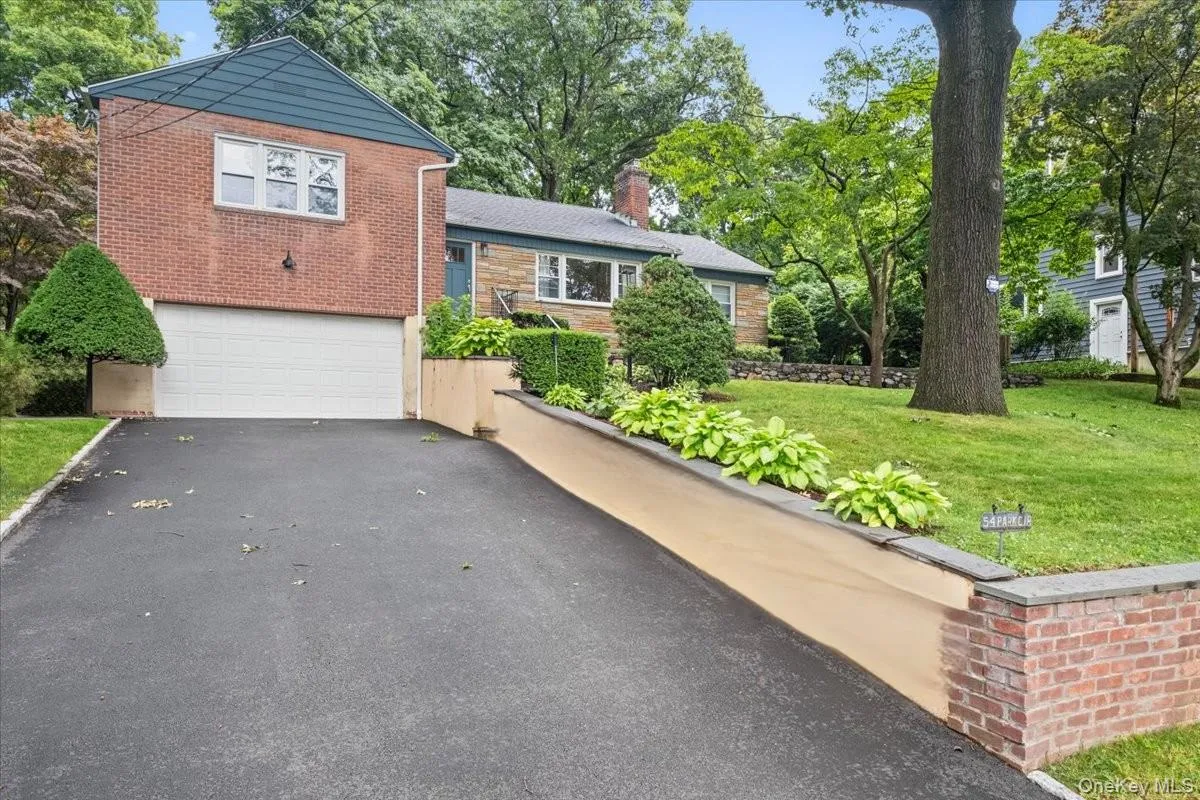 Brick facade residence featuring a two-car garage, asphalt driveway, mature tree, and landscaped retaining wall Brick facade residence featuring a two-car garage, asphalt driveway, mature tree, and landscaped retaining wall