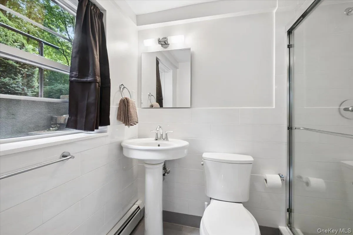 Bathroom featuring a pedestal sink, white subway tile surround, polished chrome fixtures, frameless glass shower enclosure, and a window with exterior greenery Bathroom featuring a pedestal sink, white subway tile surround, polished chrome fixtures, frameless glass shower enclosure, and a window with exterior greenery