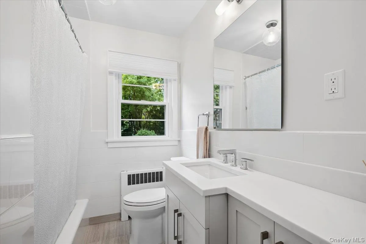 Bathroom featuring a wood-finish floor, a window with exterior foliage, a white vanity with an integrated sink, polished chrome fixtures, and a rectangular mirror Bathroom featuring a wood-finish floor, a window with exterior foliage, a white vanity with an integrated sink, polished chrome fixtures, and a rectangular mirror