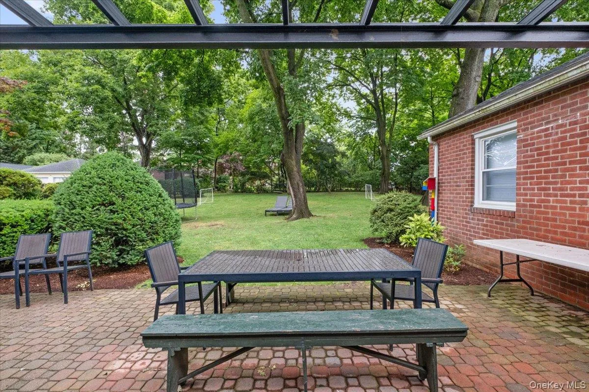 Paved patio with an open metal pergola, bordered by a brick exterior wall and established landscaping Paved patio with an open metal pergola, bordered by a brick exterior wall and established landscaping