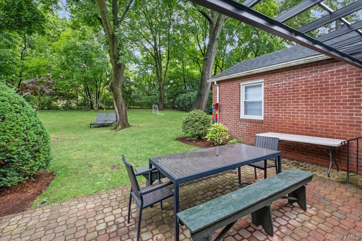 Expansive backyard featuring a brick paver patio, a partial pergola structure, lush green lawn, mature trees, and perimeter landscaping Expansive backyard featuring a brick paver patio, a partial pergola structure, lush green lawn, mature trees, and perimeter landscaping