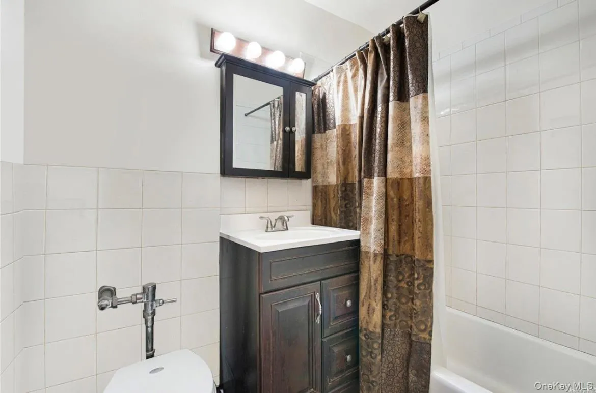 Bathroom featuring white wall tiling, a dark wood-finish vanity with a white countertop and integrated sink, a dark wood-finish medicine cabinet, and a white bathtub Bathroom featuring white wall tiling, a dark wood-finish vanity with a white countertop and integrated sink, a dark wood-finish medicine cabinet, and a white bathtub