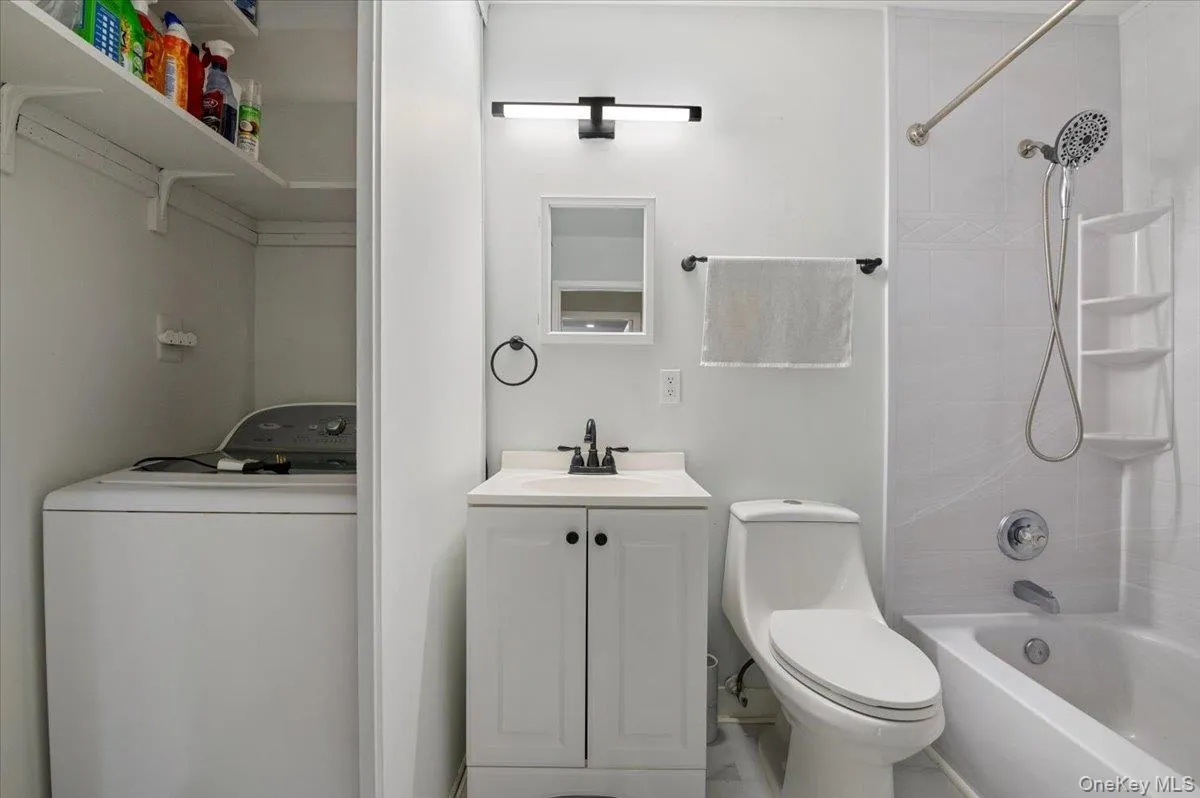 Bathroom featuring a white vanity with black hardware, a shower-over-tub with tiled walls, and a built-in laundry area with shelving Bathroom featuring a white vanity with black hardware, a shower-over-tub with tiled walls, and a built-in laundry area with shelving