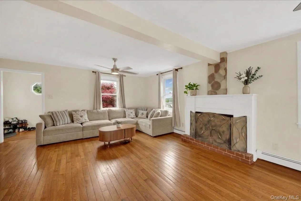 Spacious living area featuring warm wood-finish flooring, a contemporary ceiling fan, and a prominent fireplace with a white mantel and brick hearth Spacious living area featuring warm wood-finish flooring, a contemporary ceiling fan, and a prominent fireplace with a white mantel and brick hearth
