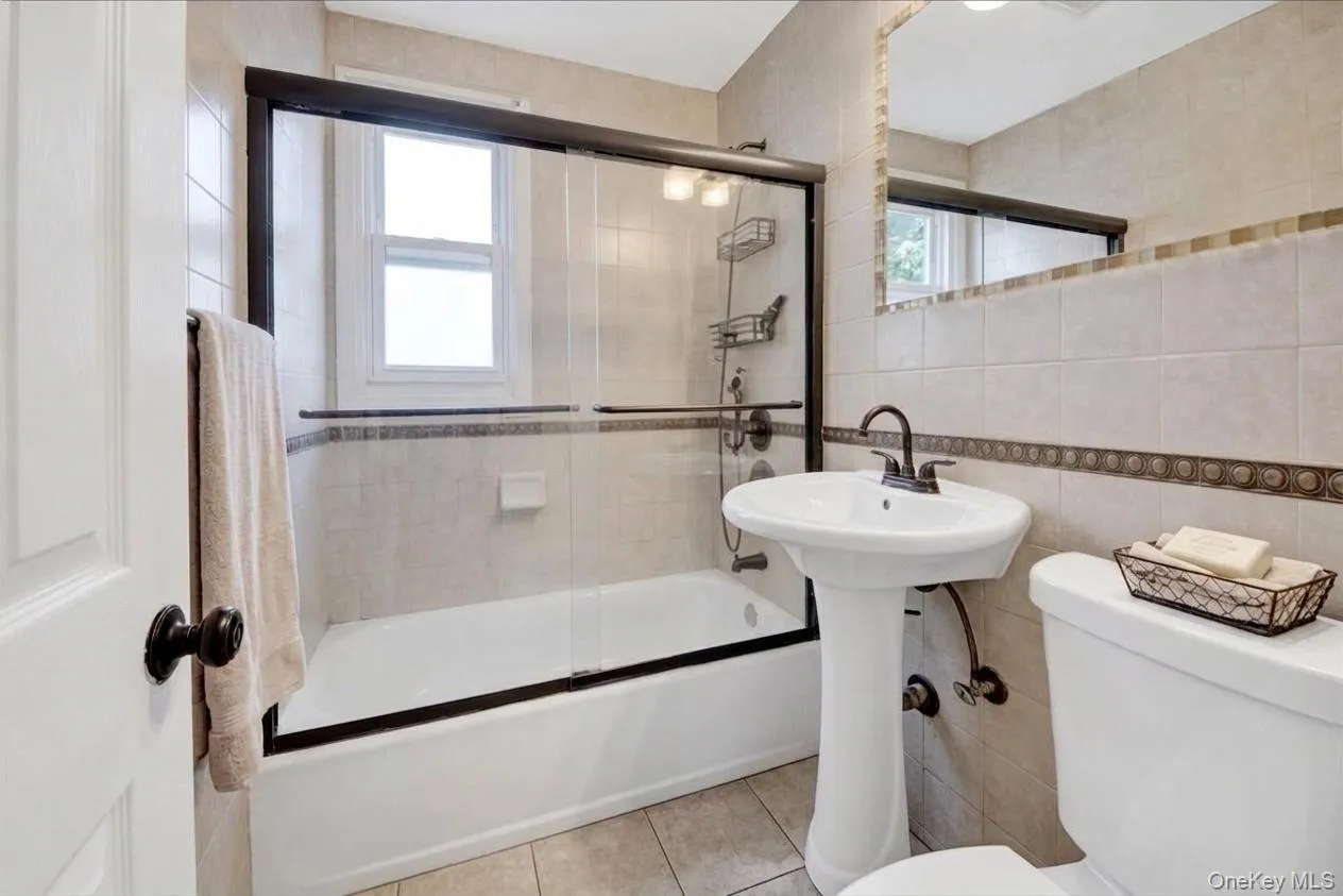 Main floor full bathroom featuring a pedestal sink with a bronze-finish faucet, a tiled shower-tub combination with sliding glass doors, and a decorative tiled accent band Main floor full bathroom featuring a pedestal sink with a bronze-finish faucet, a tiled shower-tub combination with sliding glass doors, and a decorative tiled accent band