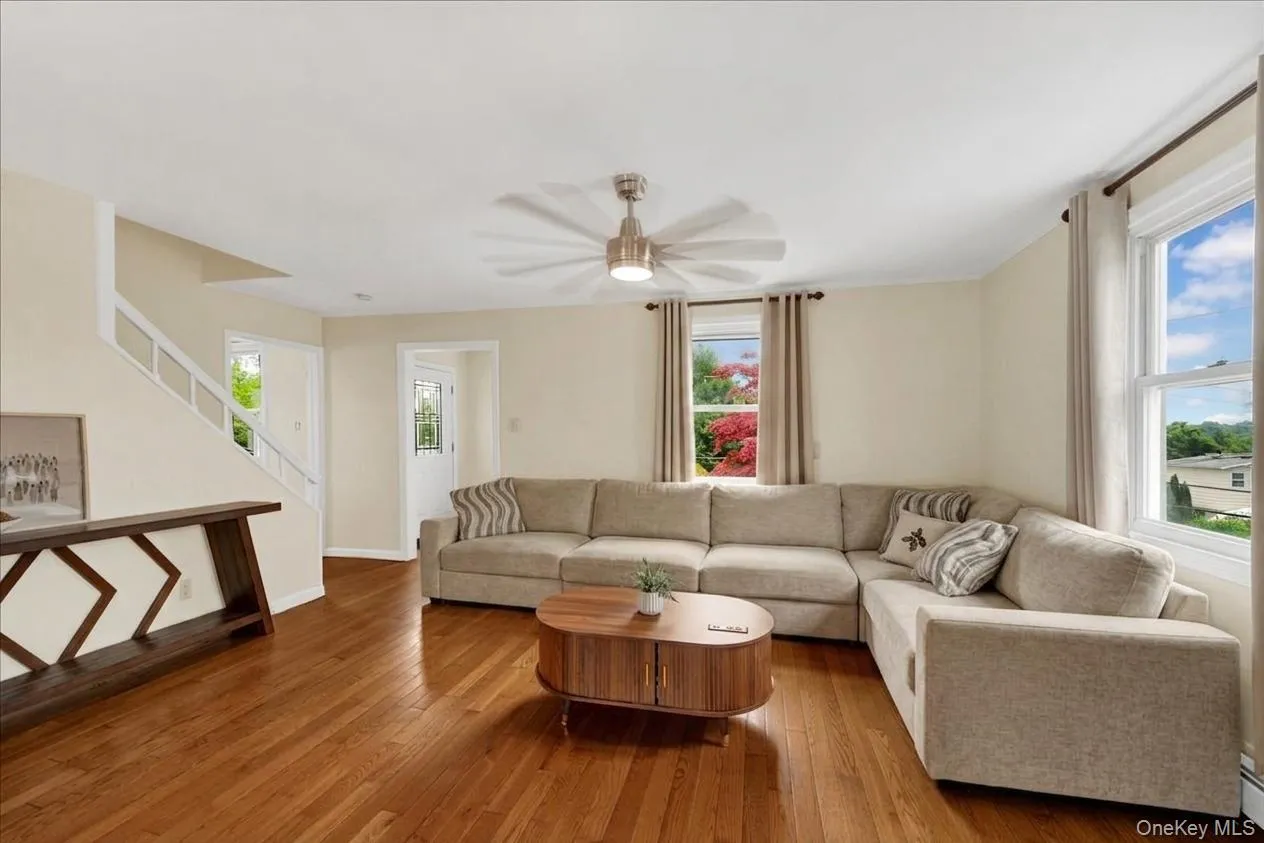 Hardwood flooring extending throughout the living space, featuring a ceiling fan, multiple windows, and a staircase with white risers and railings Hardwood flooring extending throughout the living space, featuring a ceiling fan, multiple windows, and a staircase with white risers and railings