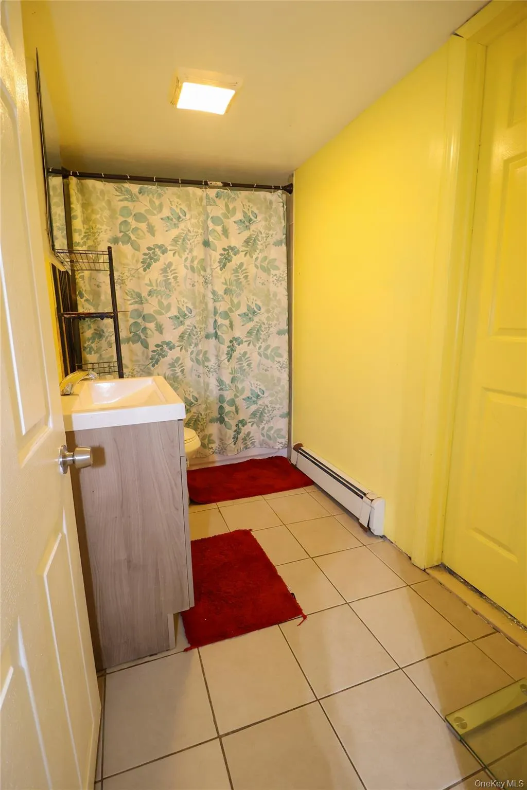 Bathroom featuring light-toned tile flooring, a shower area with a curtain, a vanity with a light wood-finish cabinet and white countertop, and a wall-mounted baseboard heater Bathroom featuring light-toned tile flooring, a shower area with a curtain, a vanity with a light wood-finish cabinet and white countertop, and a wall-mounted baseboard heater