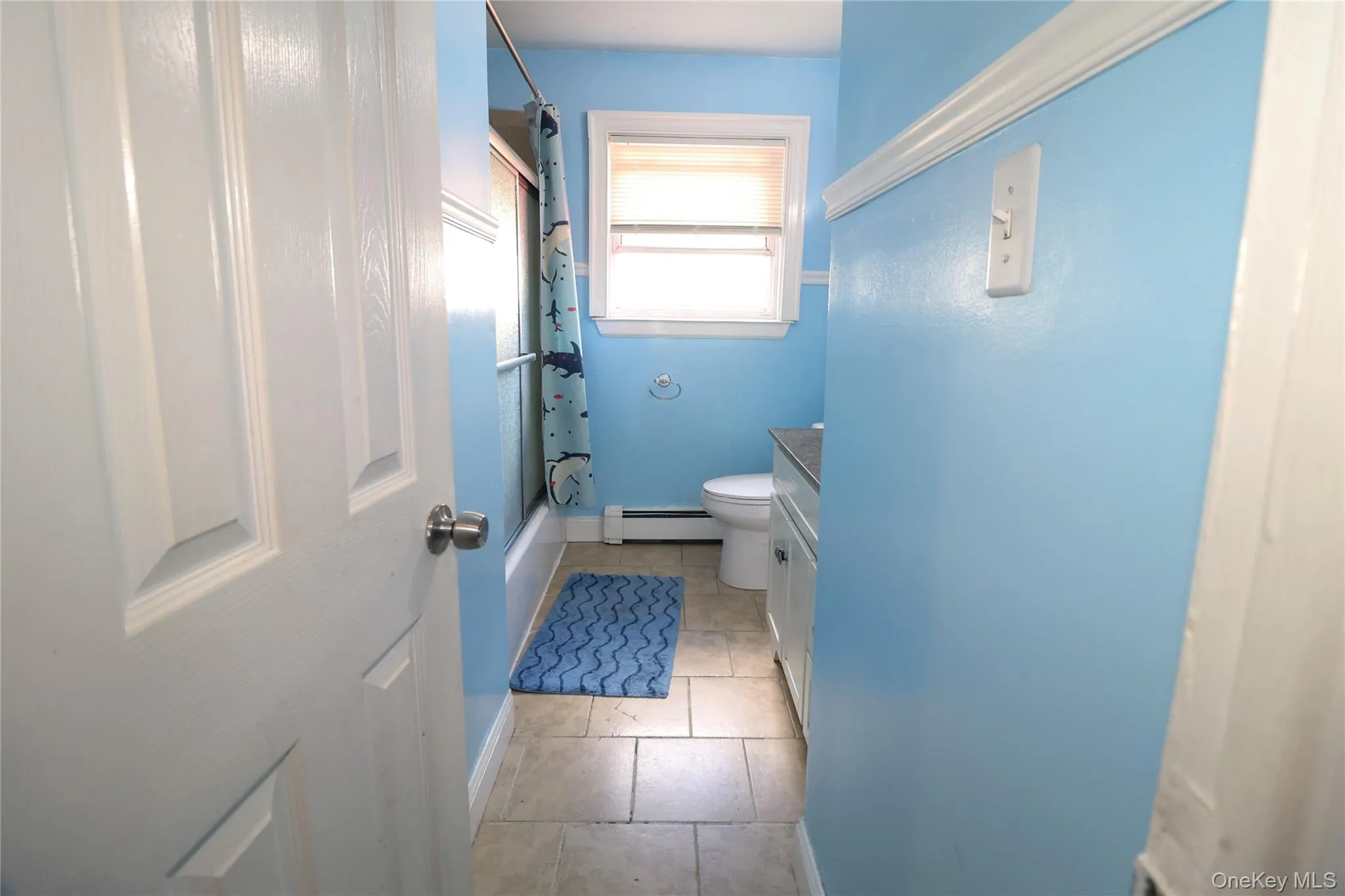 Bathroom featuring light blue walls, tiled flooring, and a single window with blinds Bathroom featuring light blue walls, tiled flooring, and a single window with blinds