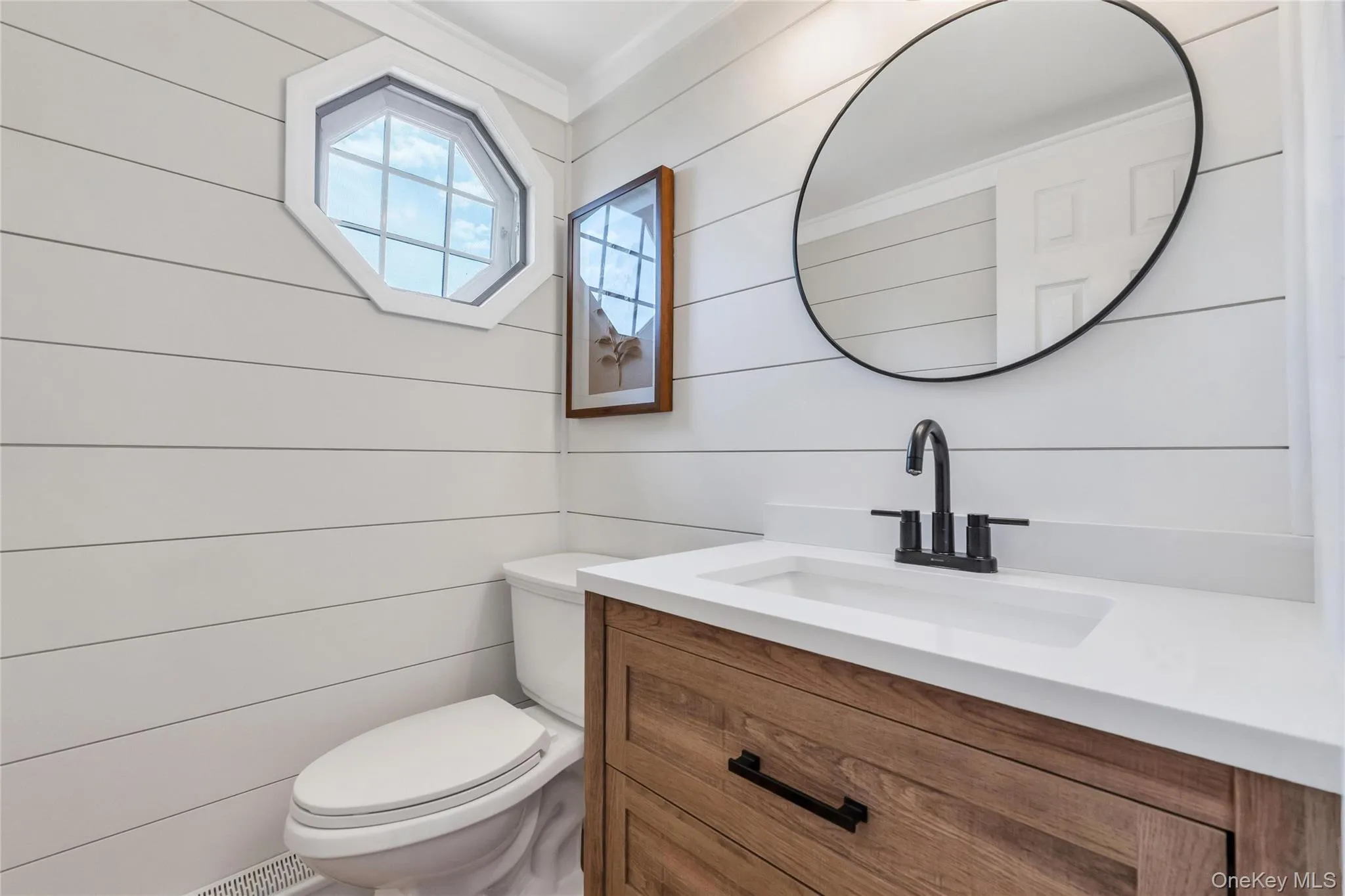 Bathroom featuring shiplap walls, an octagon window, and a wood-finish vanity with a white countertop and matte black hardware Bathroom featuring shiplap walls, an octagon window, and a wood-finish vanity with a white countertop and matte black hardware