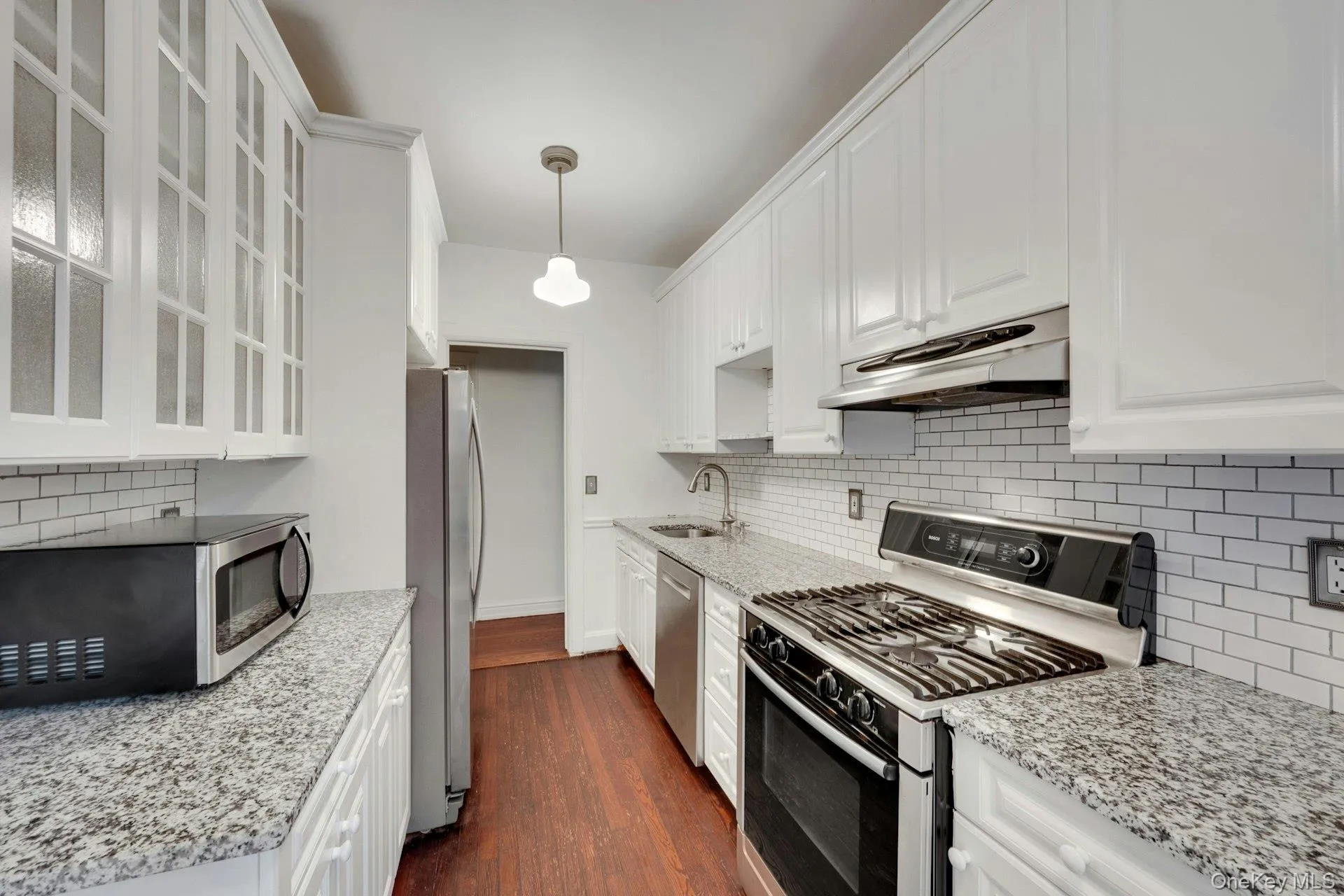 Galley kitchen featuring white cabinetry, light granite countertops, stainless steel appliances, a white subway tile backsplash, and wood-finish flooring Galley kitchen featuring white cabinetry, light granite countertops, stainless steel appliances, a white subway tile backsplash, and wood-finish flooring