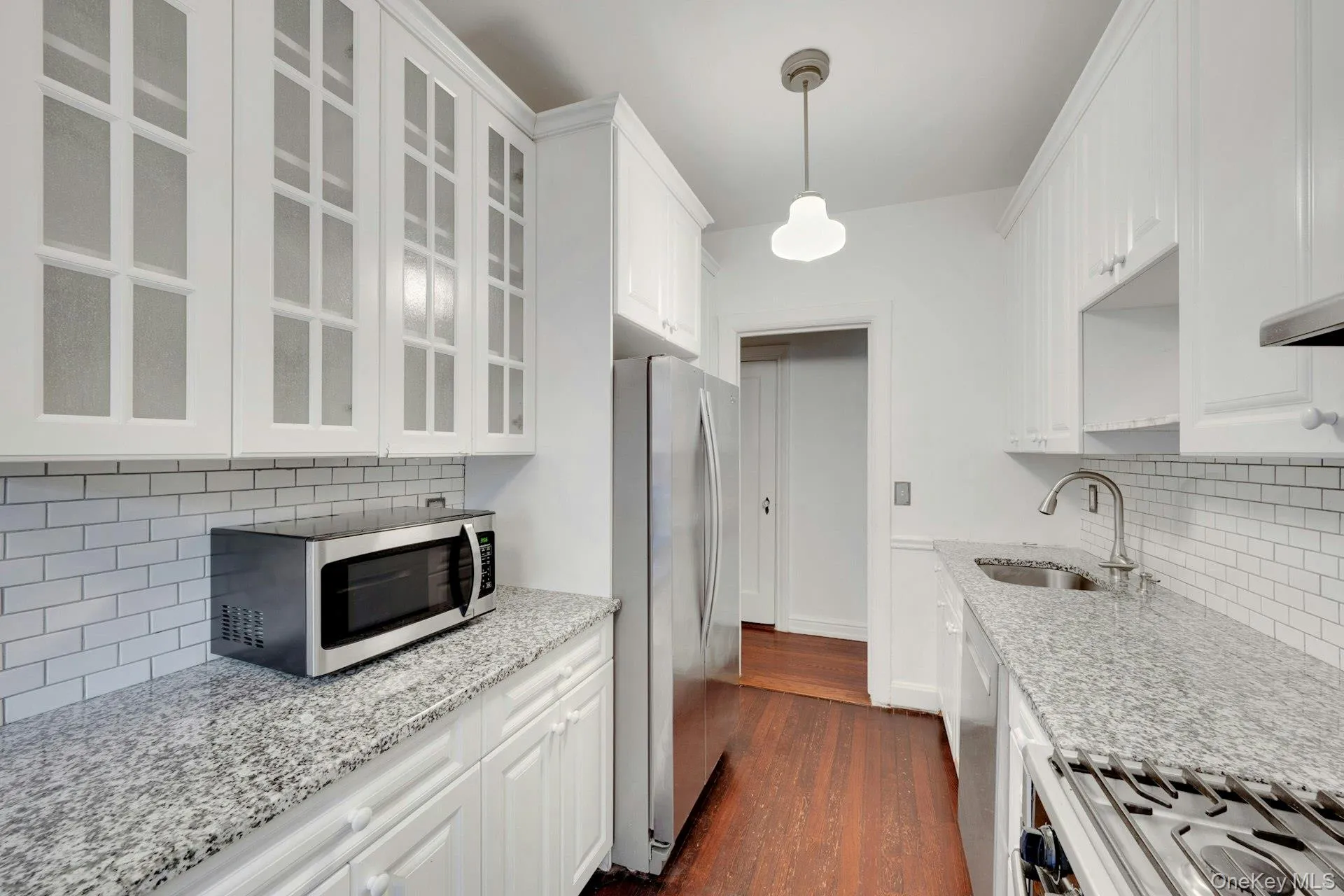 Galley kitchen featuring white cabinetry, glass-front upper cabinets, granite countertops, and a subway tile backsplash Galley kitchen featuring white cabinetry, glass-front upper cabinets, granite countertops, and a subway tile backsplash