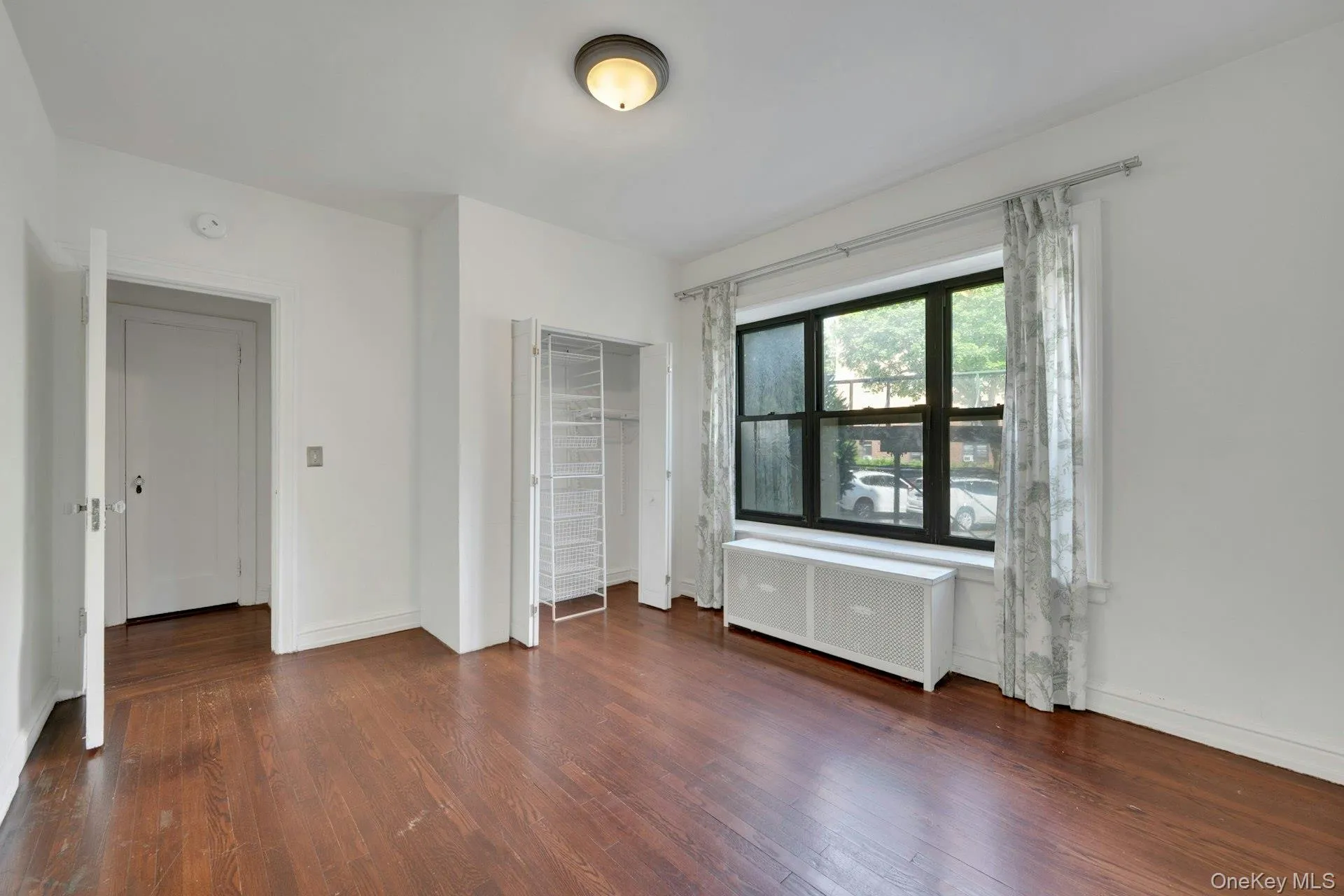 Spacious room featuring wood-finish flooring, a large multi-pane window, and a built-in radiator cover Spacious room featuring wood-finish flooring, a large multi-pane window, and a built-in radiator cover