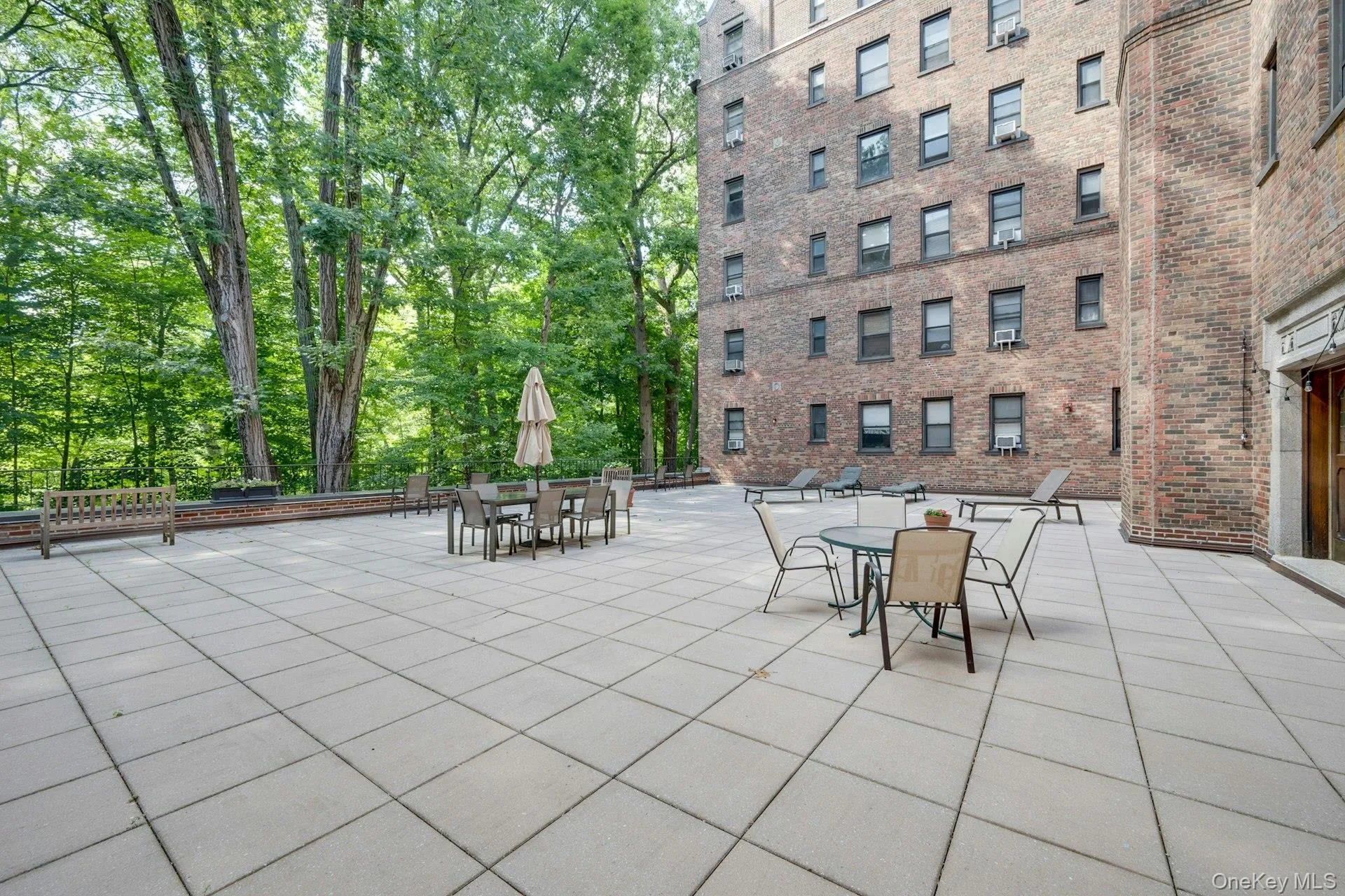 Expansive outdoor patio featuring large square pavers, a low brick retaining wall, and mature trees providing a natural backdrop Expansive outdoor patio featuring large square pavers, a low brick retaining wall, and mature trees providing a natural backdrop