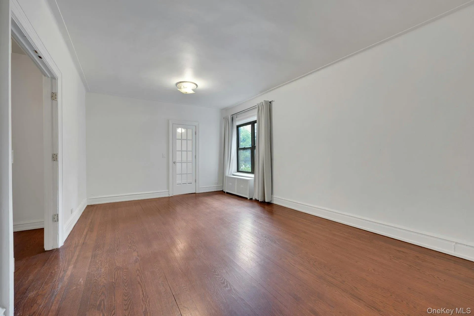 Hardwood flooring present in an expansive room featuring a multi-pane glass door, a window with drapery, and a surface-mounted ceiling light fixture Hardwood flooring present in an expansive room featuring a multi-pane glass door, a window with drapery, and a surface-mounted ceiling light fixture
