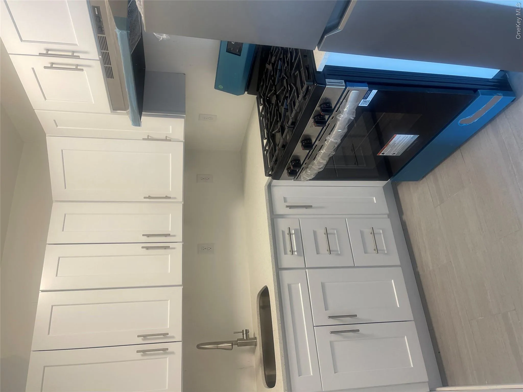 Modern kitchen featuring white shaker cabinetry with brushed nickel hardware, light-toned countertops, a stainless steel sink with a gooseneck faucet, a full-size range, and light grey tile flooring Modern kitchen featuring white shaker cabinetry with brushed nickel hardware, light-toned countertops, a stainless steel sink with a gooseneck faucet, a full-size range, and light grey tile flooring
