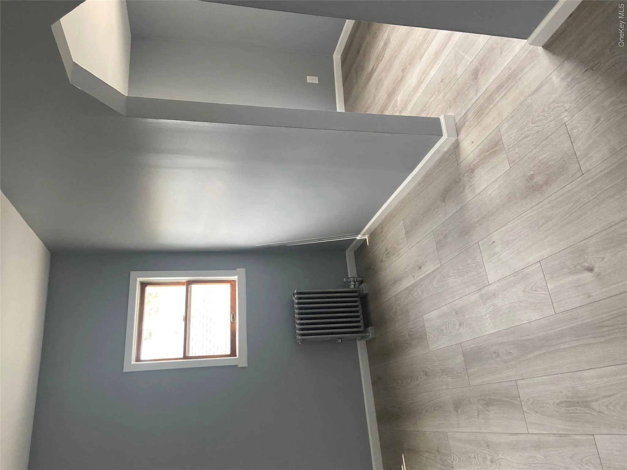 Light wood-finish flooring extends throughout the space, complemented by a radiator beneath a window with white trim Light wood-finish flooring extends throughout the space, complemented by a radiator beneath a window with white trim