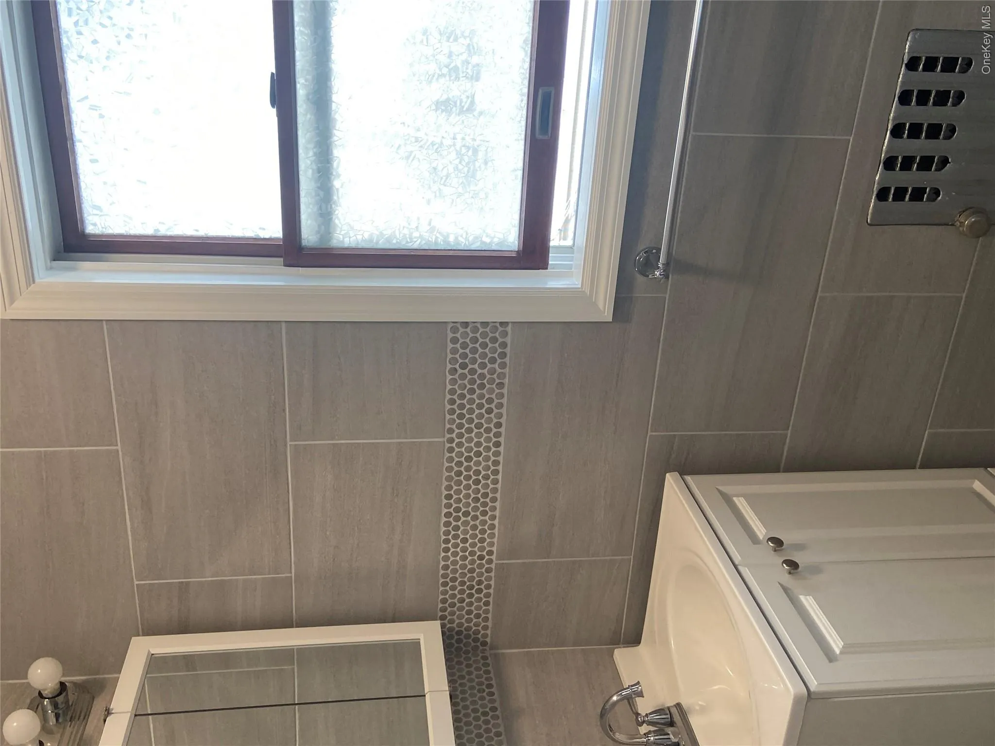 Bathroom featuring large format gray wall tiling with a mosaic accent band, a white vanity with integrated sink, chrome faucet, and a window with frosted glass Bathroom featuring large format gray wall tiling with a mosaic accent band, a white vanity with integrated sink, chrome faucet, and a window with frosted glass