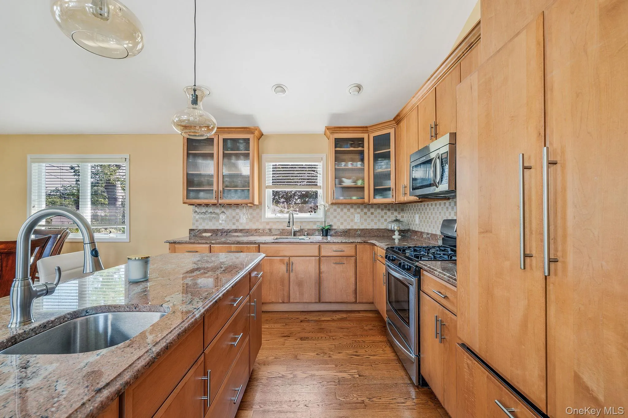Kitchen with light wood-style floors, appliances with stainless steel finishes, glass insert cabinets, light stone countertops, and decorative light fixtures Kitchen with light wood-style floors, appliances with stainless steel finishes, glass insert cabinets, light stone countertops, and decorative light fixtures