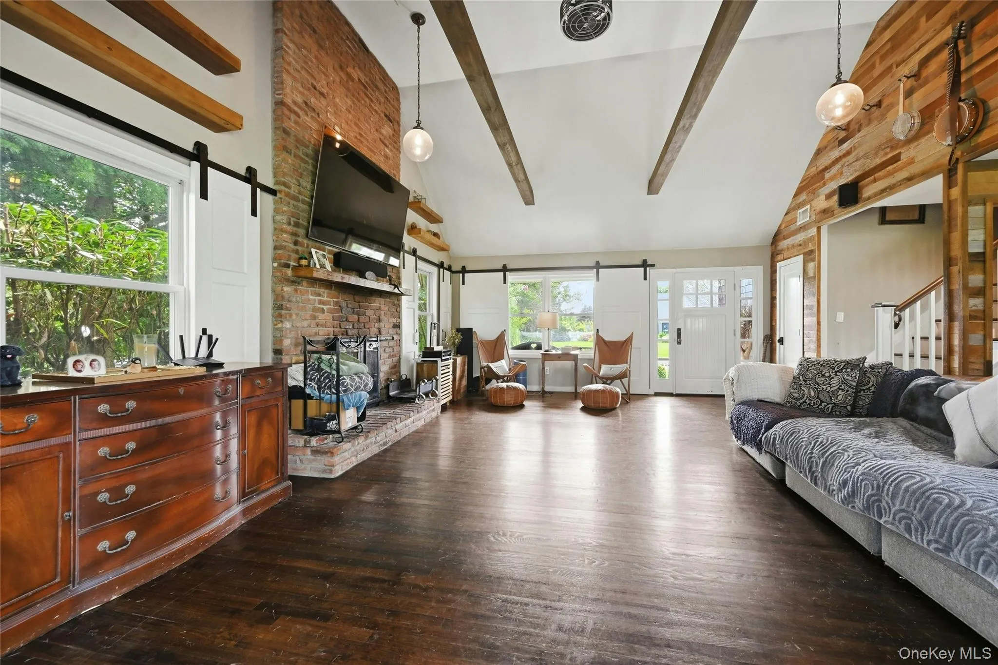 Spacious living area featuring exposed ceiling beams, a brick fireplace, wood-finish flooring, and a front entry door with side windows Spacious living area featuring exposed ceiling beams, a brick fireplace, wood-finish flooring, and a front entry door with side windows