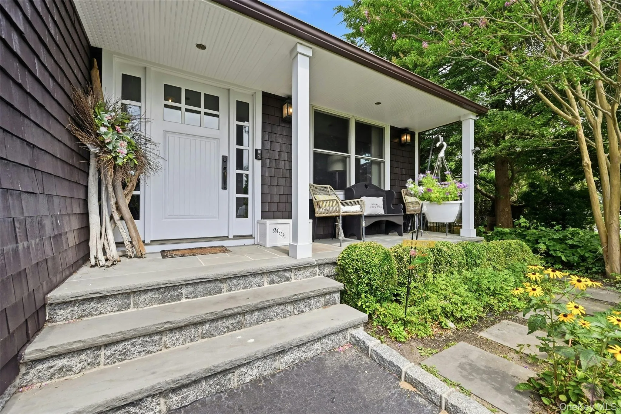 Welcoming front entry featuring a white paneled door with sidelights, a covered porch with white columns, stone steps, dark-stained cedar shake siding, and mature landscaping Welcoming front entry featuring a white paneled door with sidelights, a covered porch with white columns, stone steps, dark-stained cedar shake siding, and mature landscaping
