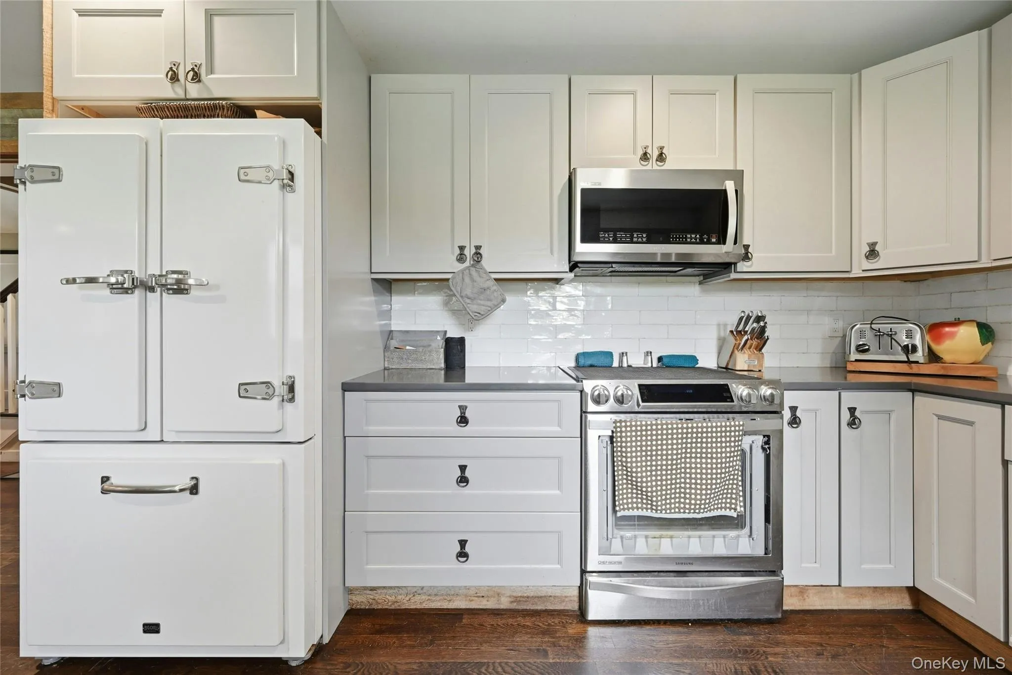 Kitchen featuring stainless steel appliances, white shaker-style cabinetry, dark countertops, a white subway tile backsplash, and wood-finish flooring Kitchen featuring stainless steel appliances, white shaker-style cabinetry, dark countertops, a white subway tile backsplash, and wood-finish flooring