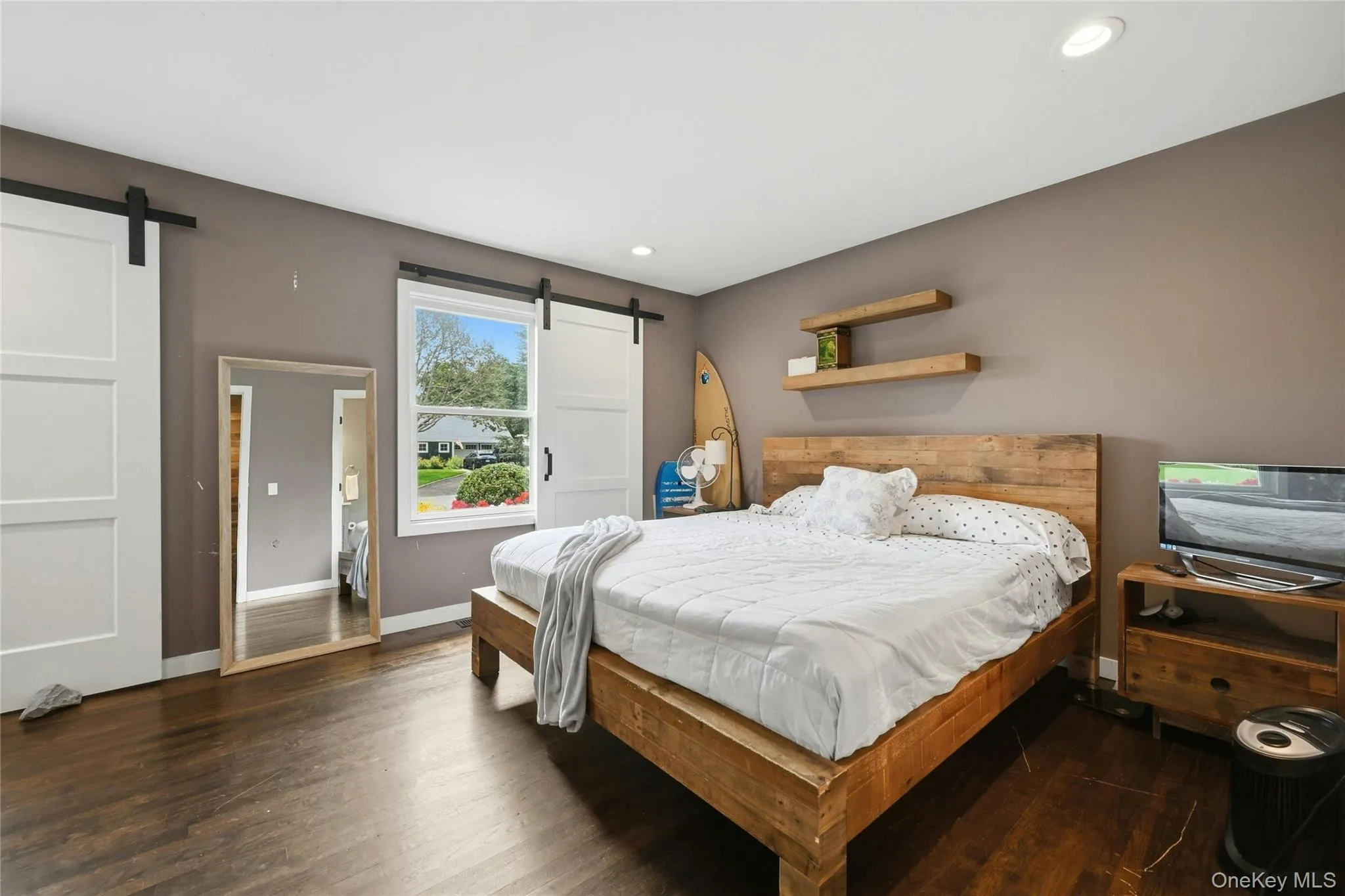 Spacious primary bed-room featuring dark wood-finish flooring, recessed lighting, and a window with white trim, plantation style shutter as window-treatment. Spacious primary bed-room featuring dark wood-finish flooring, recessed lighting, and a window with white trim, plantation style shutter as window-treatment.