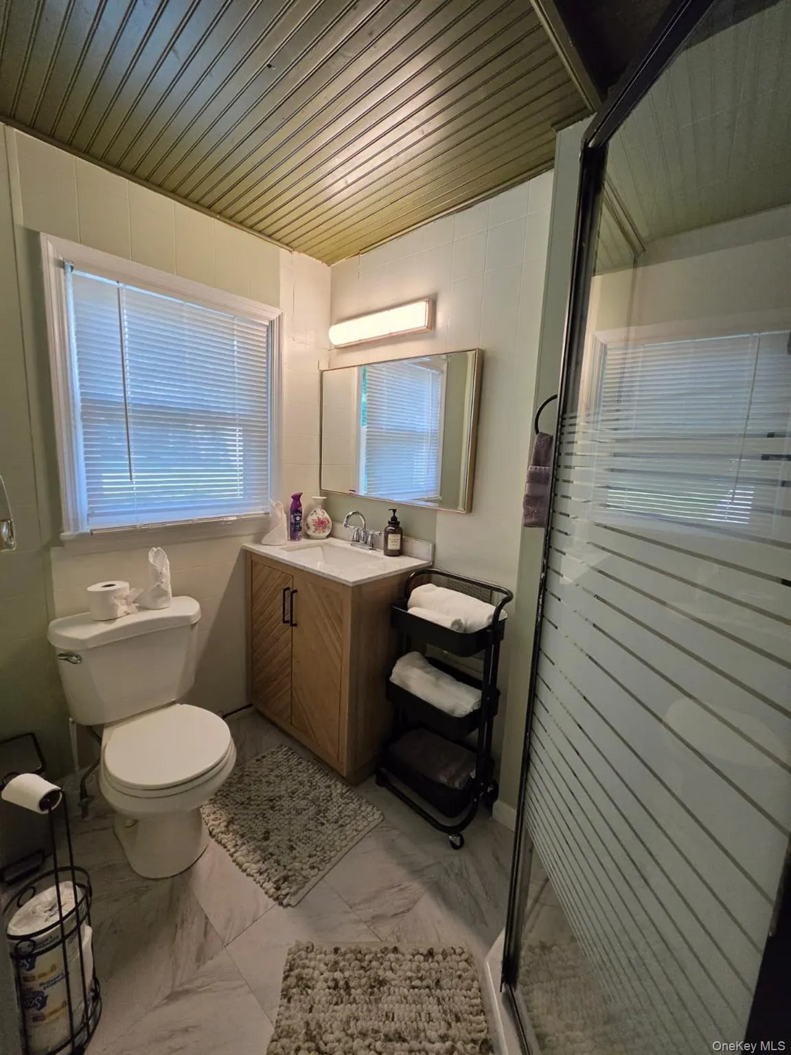 Main Floor Bathroom featuring a walk-in shower with frosted glass enclosure, a single vanity with wood-finish cabinetry, a rectangular sink with chrome faucet, light-toned wall paneling, and a wood-slat ceiling Main Floor Bathroom featuring a walk-in shower with frosted glass enclosure, a single vanity with wood-finish cabinetry, a rectangular sink with chrome faucet, light-toned wall paneling, and a wood-slat ceiling