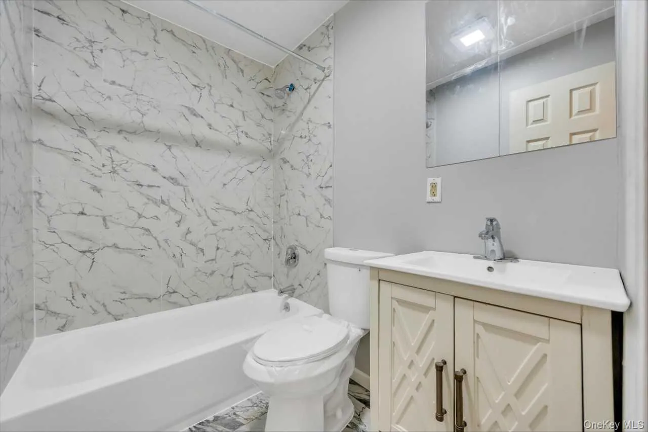 Bathroom featuring a white bathtub, marble-look tiled shower surround, light grey painted walls, a white vanity with decorative cabinet doors, and a mirrored medicine cabinet Bathroom featuring a white bathtub, marble-look tiled shower surround, light grey painted walls, a white vanity with decorative cabinet doors, and a mirrored medicine cabinet