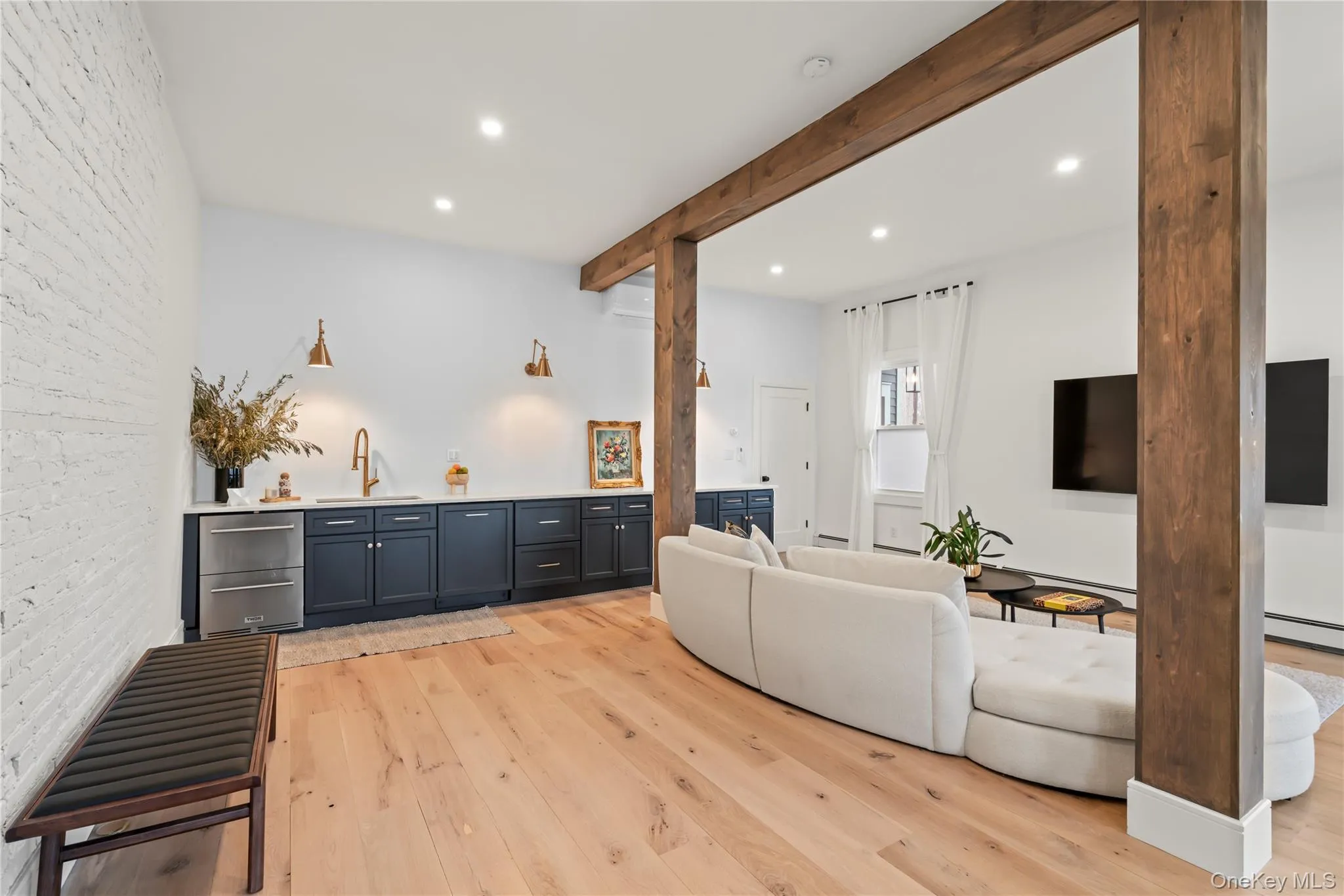 LowerOpen-concept living area featuring exposed wood beams, light wood-finish flooring, and a white brick accent wall LowerOpen-concept living area featuring exposed wood beams, light wood-finish flooring, and a white brick accent wall