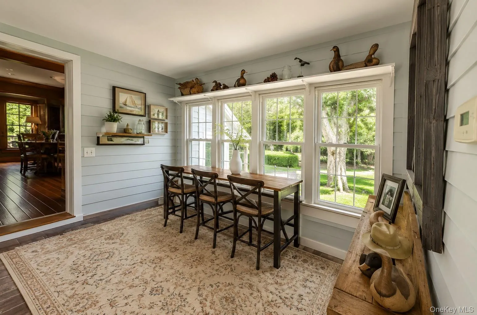 Sunlit interior space featuring multiple large windows, light blue shiplap accent walls, and a wide entryway to an additional room with wood-finish flooring Sunlit interior space featuring multiple large windows, light blue shiplap accent walls, and a wide entryway to an additional room with wood-finish flooring