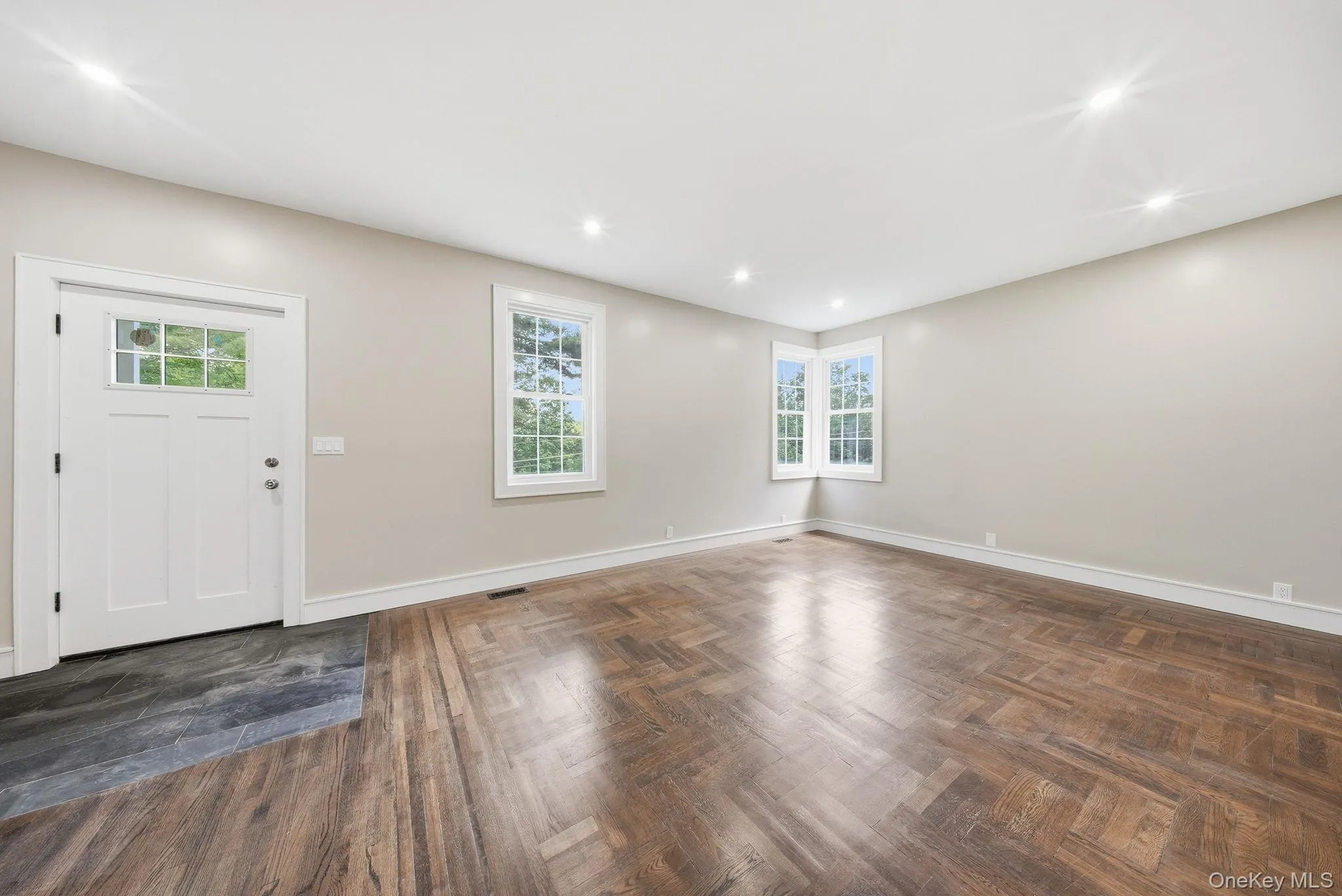 Entry foyer and living area featuring herringbone wood-finish flooring, recessed lighting, a paneled entry door with transom window, and a corner window arrangement Entry foyer and living area featuring herringbone wood-finish flooring, recessed lighting, a paneled entry door with transom window, and a corner window arrangement