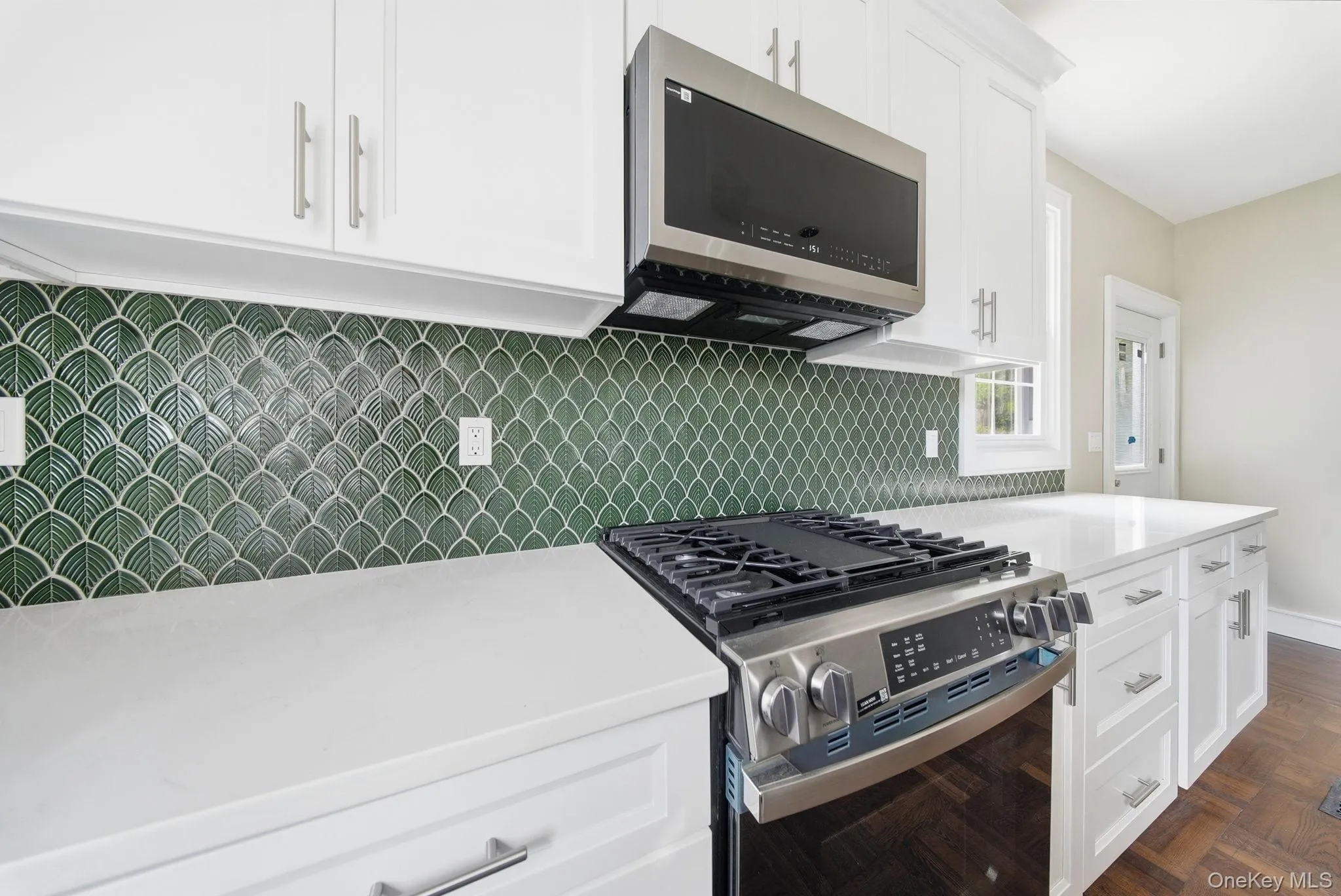 Kitchen featuring white cabinetry, light-toned countertops, a stainless steel range with overhead microwave, and a distinctive green scallop-patterned tile backsplash Kitchen featuring white cabinetry, light-toned countertops, a stainless steel range with overhead microwave, and a distinctive green scallop-patterned tile backsplash