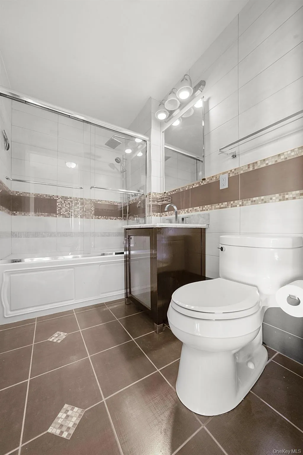 Bathroom featuring a built-in bathtub with glass sliding doors, a vanity with a dark wood-finish base, a rectangular mirror with integrated track lighting, and patterned floor tiling Bathroom featuring a built-in bathtub with glass sliding doors, a vanity with a dark wood-finish base, a rectangular mirror with integrated track lighting, and patterned floor tiling